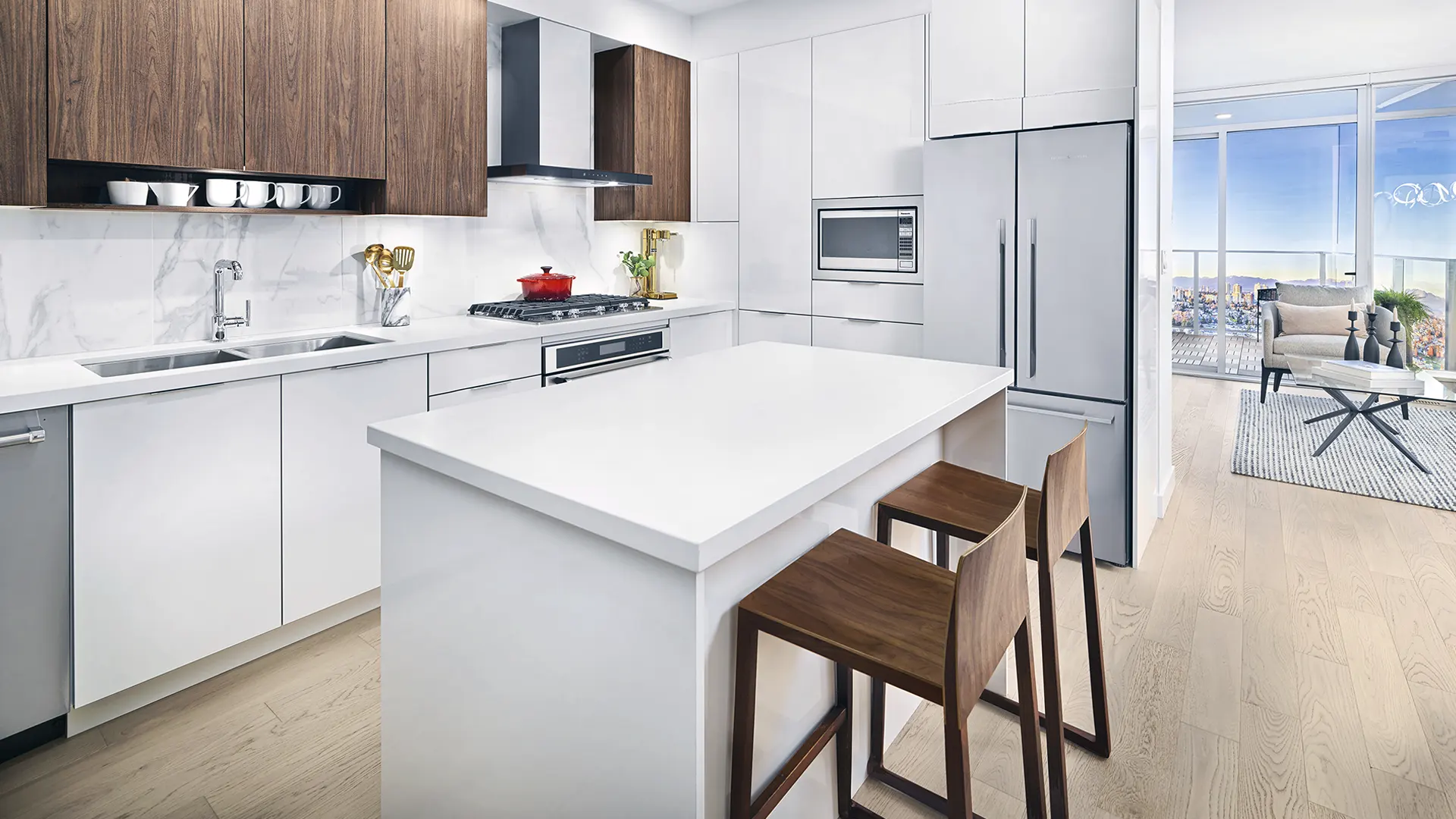 Southgate City – A kitchen with white cabinetry, dark wood upper cabinets, a stainless steel french-door refrigerator, a large white island with bar stools, and a view through to a living room and balcony with a city view.