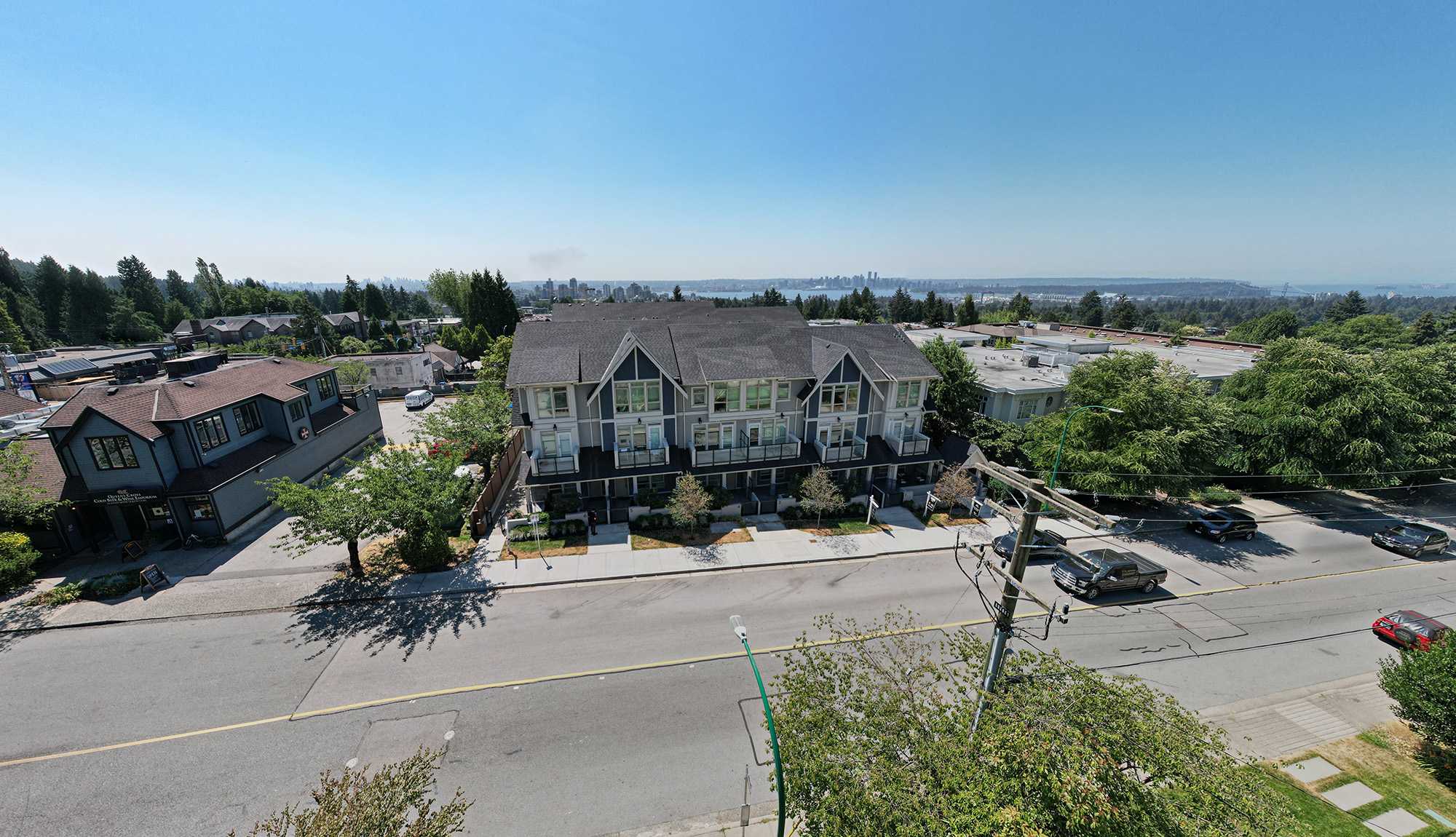 Queen’s Landing – An elevated drone shot shows a modern grey three-story townhome complex situated along a suburban street under a clear blue sky. In the background, green treetops stretch toward a distant water body and city skyline under a bright, sunny daytime light.