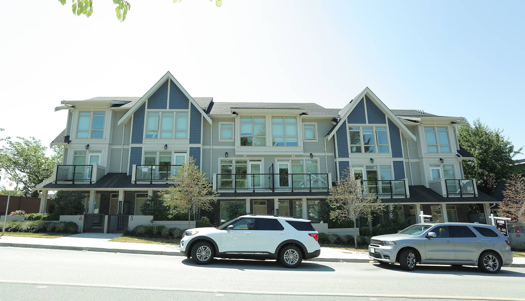 Queen’s Landing – A ground-level view of a modern three-story townhouse complex featuring blue and light grey siding, prominent gables, and private upper-floor balconies with glass railings. Two SUVs are parked along the paved street in front of the landscaped front entrance on a bright, clear day.