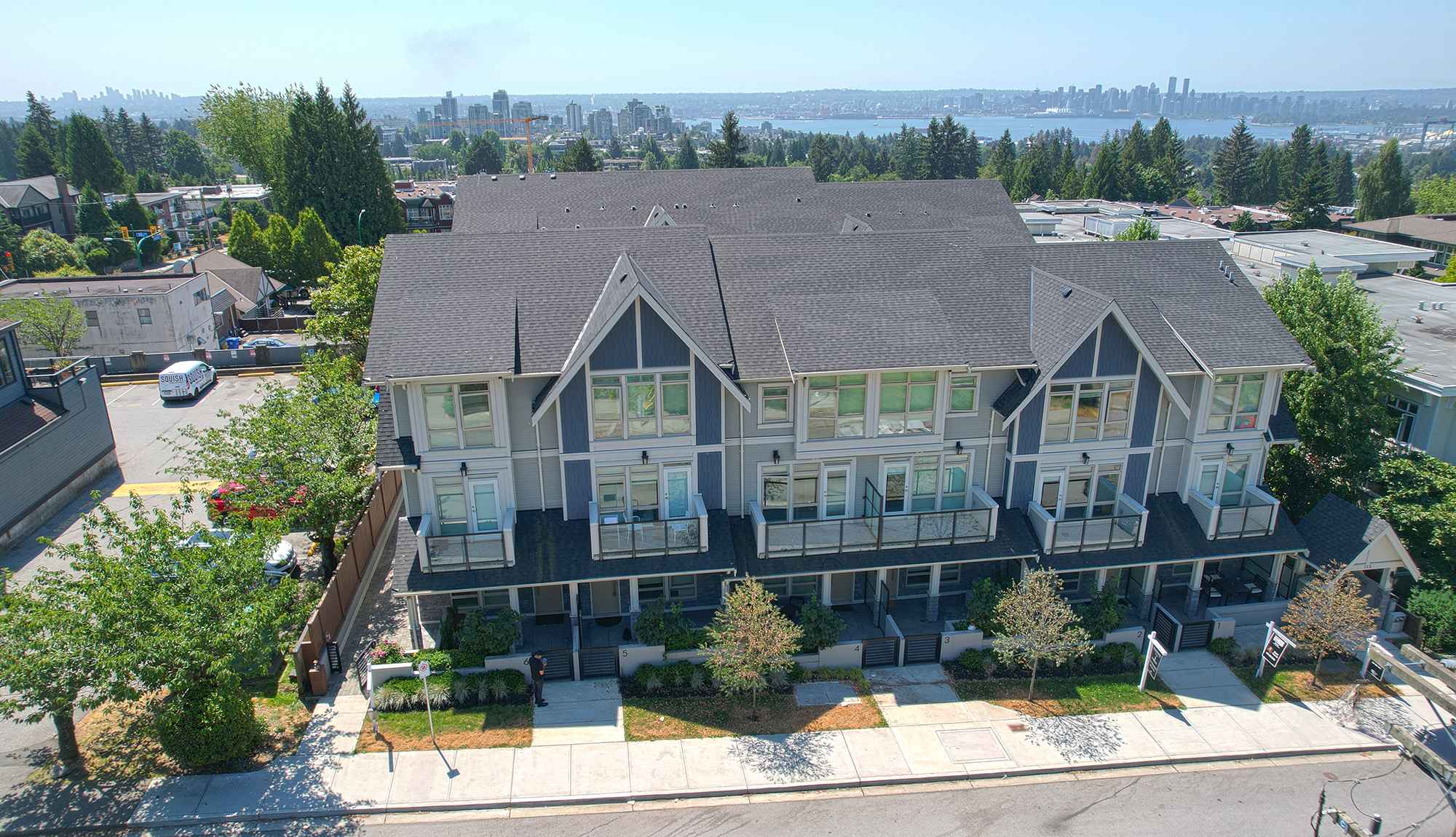 Queen’s Landing – An elevated aerial view captures a multi-story, craftsman-style townhouse complex with dark roofs, blue and grey paneling, and glass balconies under a bright, clear sky. In the background, lush green treetops extend toward a body of water and a distant city skyline.