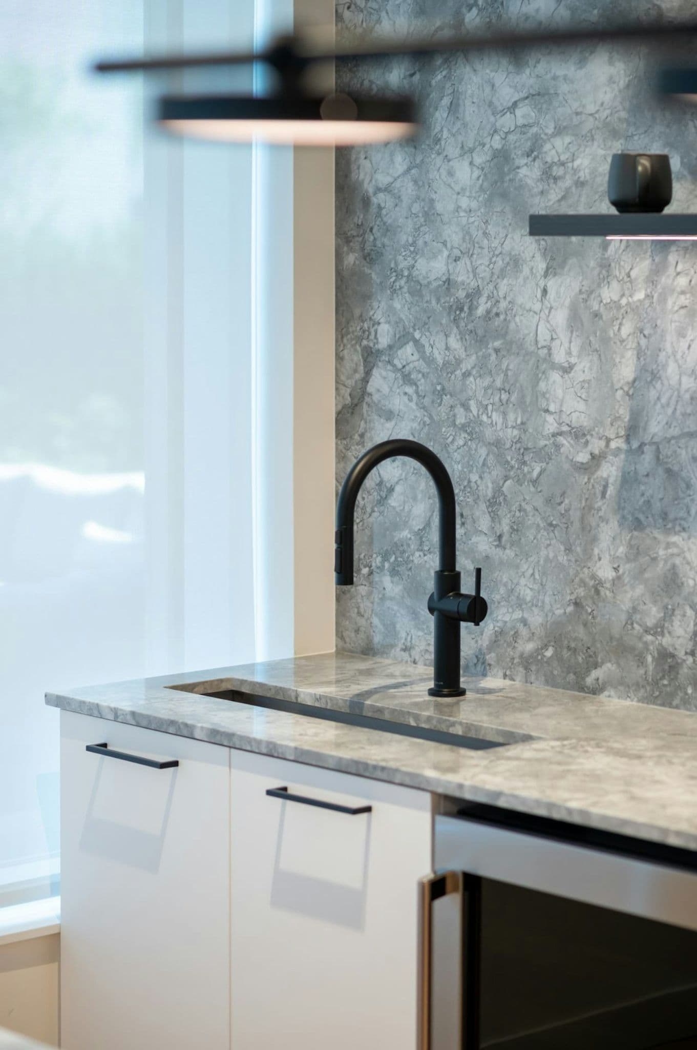 The Standard – Close-up of a kitchen island sink area with a matte black faucet and gray marble-look countertop and backsplash.
