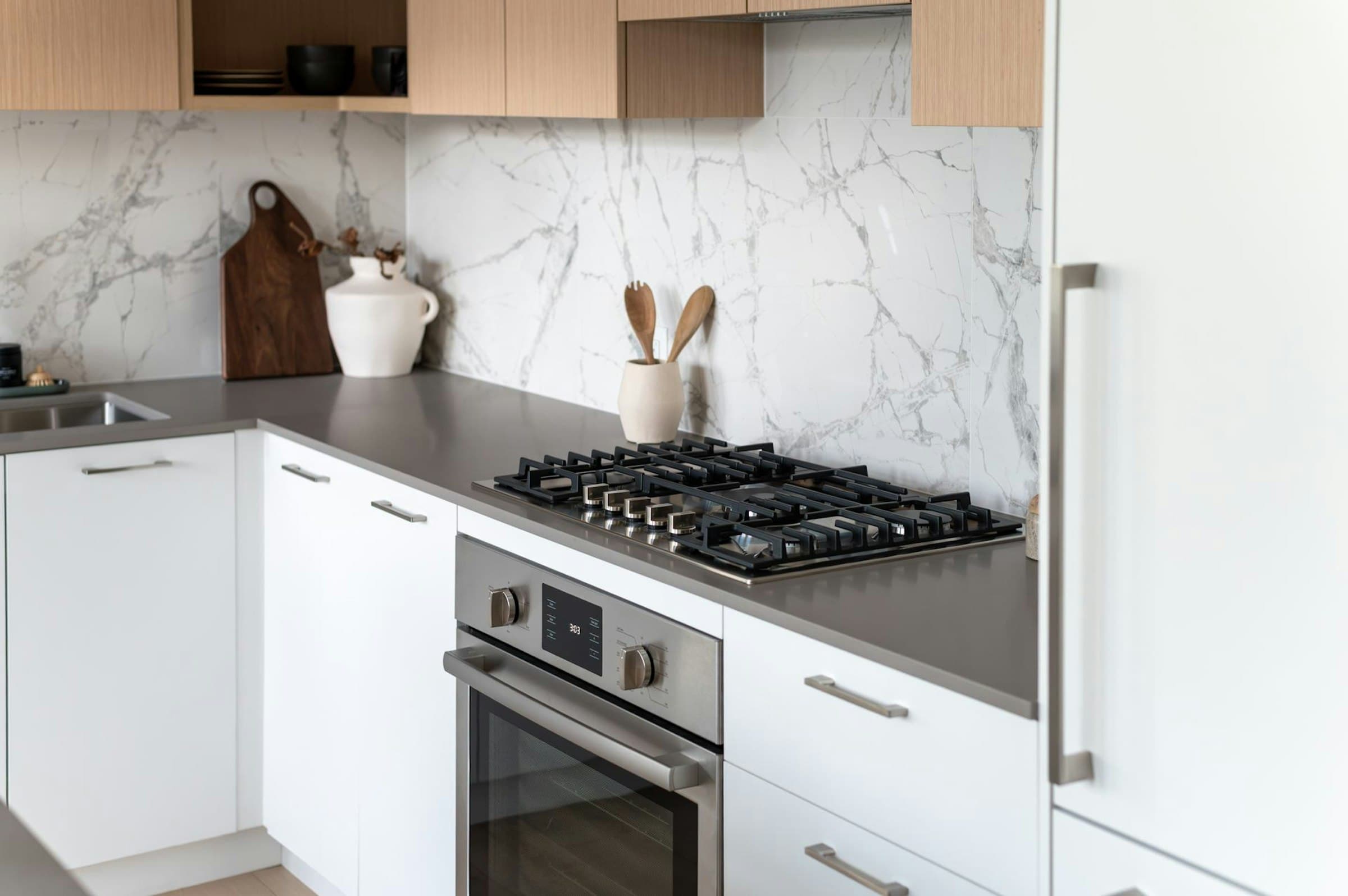 The Standard – Close-up of a kitchen with light wood upper cabinets, marble-look backsplash, gas cooktop, and built-in wall oven.