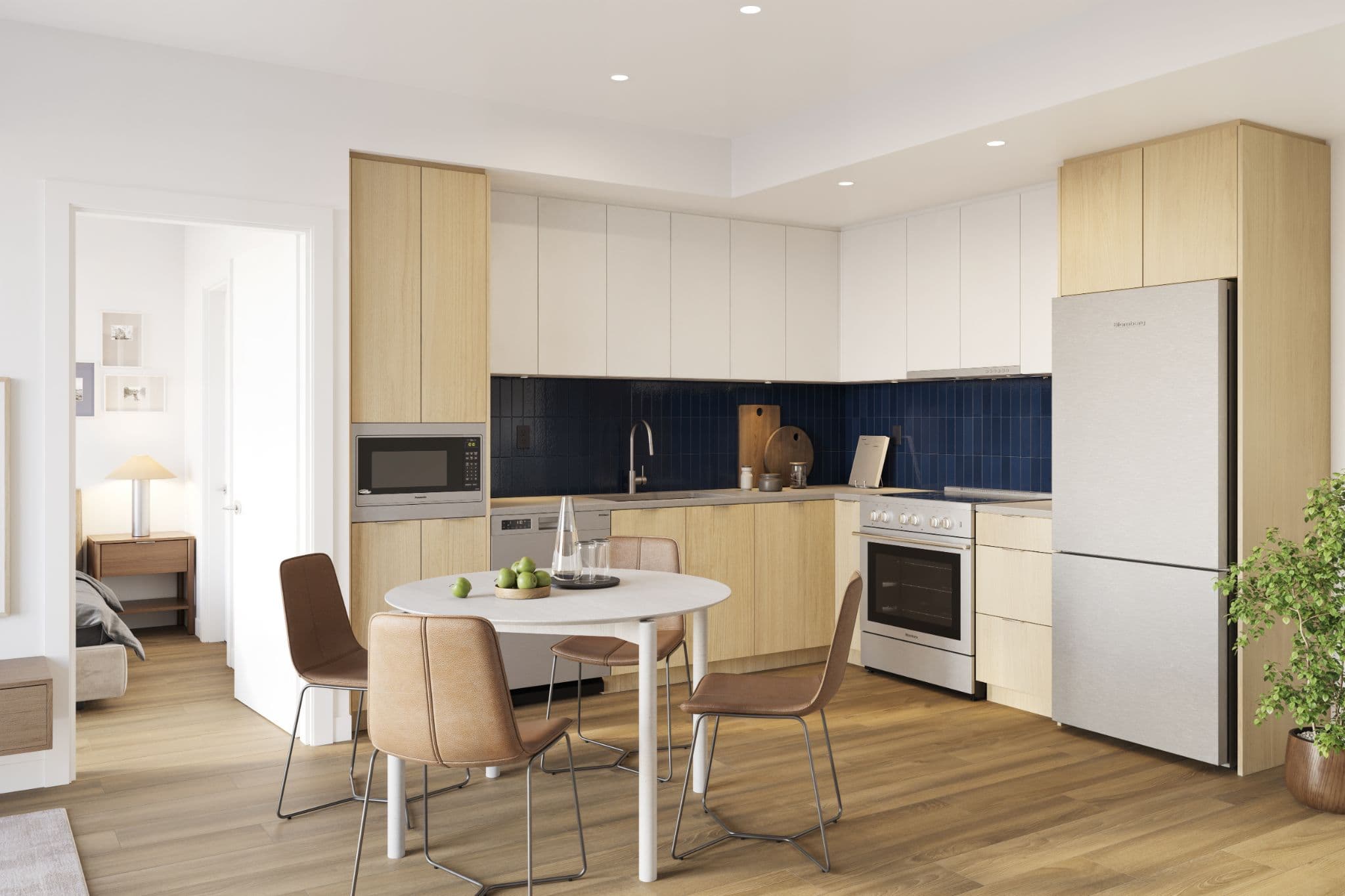 Market Commons – A modern open-concept apartment kitchen and dining area featuring two-tone cabinetry with light wood lower cabinets, white upper cabinets, stainless steel appliances, and a deep blue tiled backsplash. In the foreground sits a small round dining table with four brown leather chairs on light hardwood floors, with a doorway offering a glimpse into an adjacent bedroom.