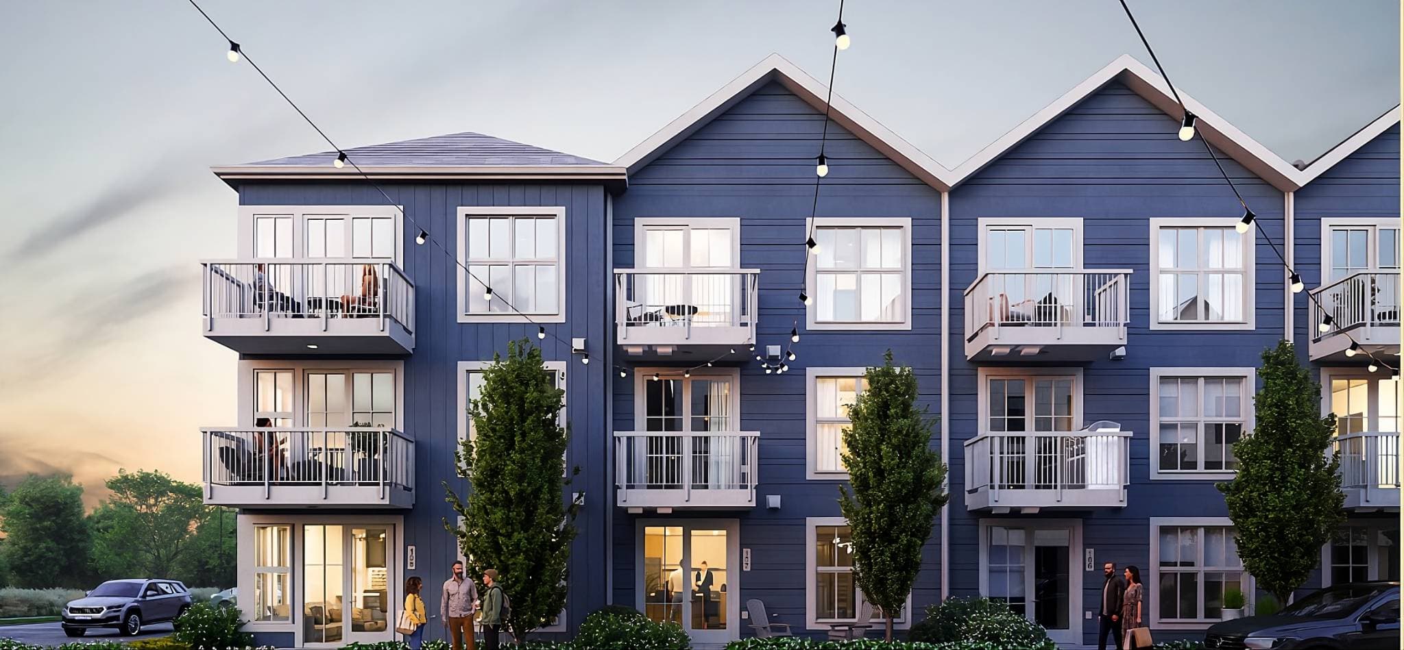 Market Commons – A dusk view of a three-story, blue siding townhome development with pitched rooflines, white trim, and individual balconies. Overhead string lights crisscross in front of the building, illuminating residents walking outside and warm interior lights visible through large glass doors and windows.