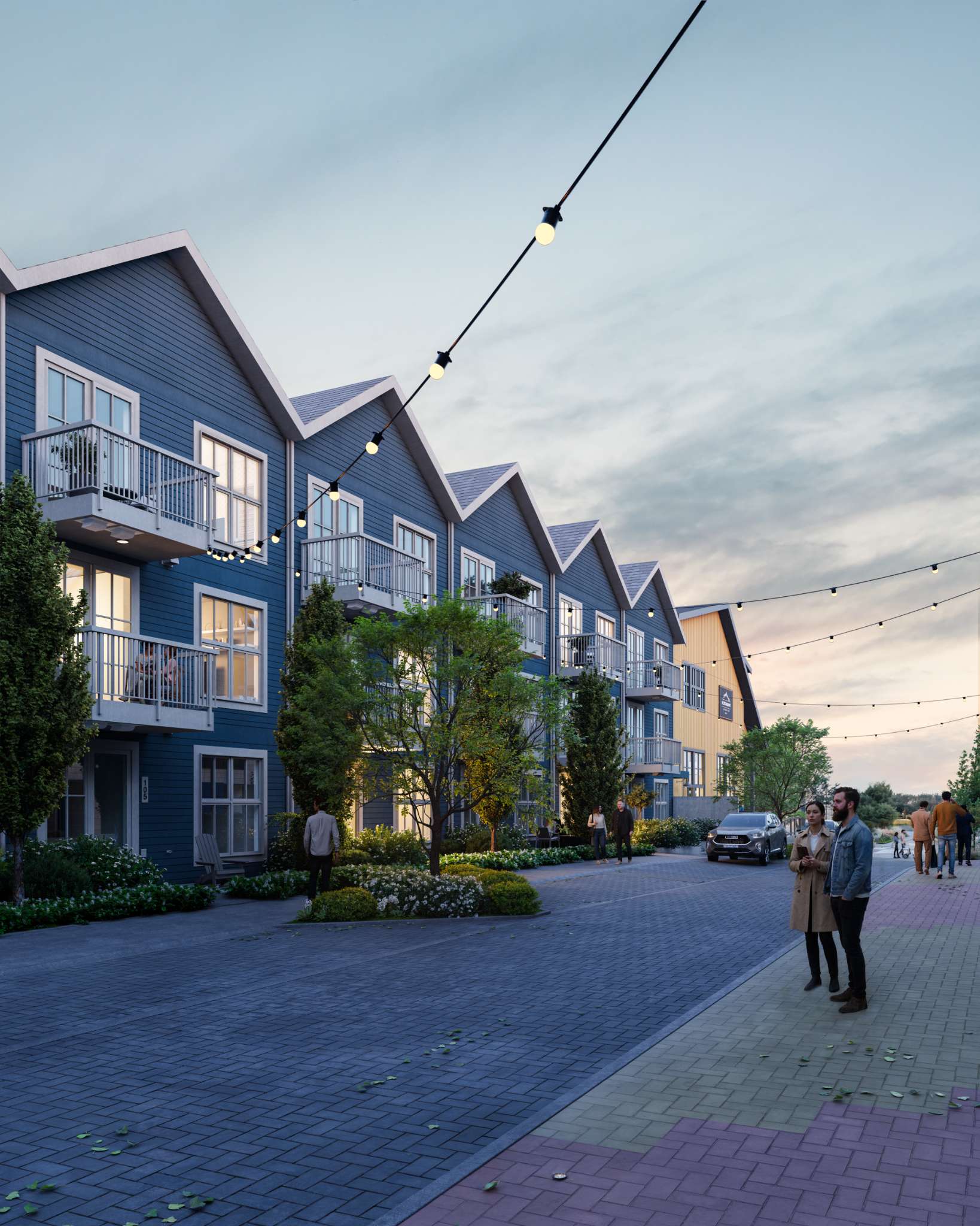 Market Commons – A row of multi-story gabled residential townhomes with blue siding and private balconies lines a paved pedestrian-friendly street adorned with overhead string lights. In the dusk setting, warm light spills from interior windows while residents stroll along the sidewalks beside landscaped flowerbeds and young trees.