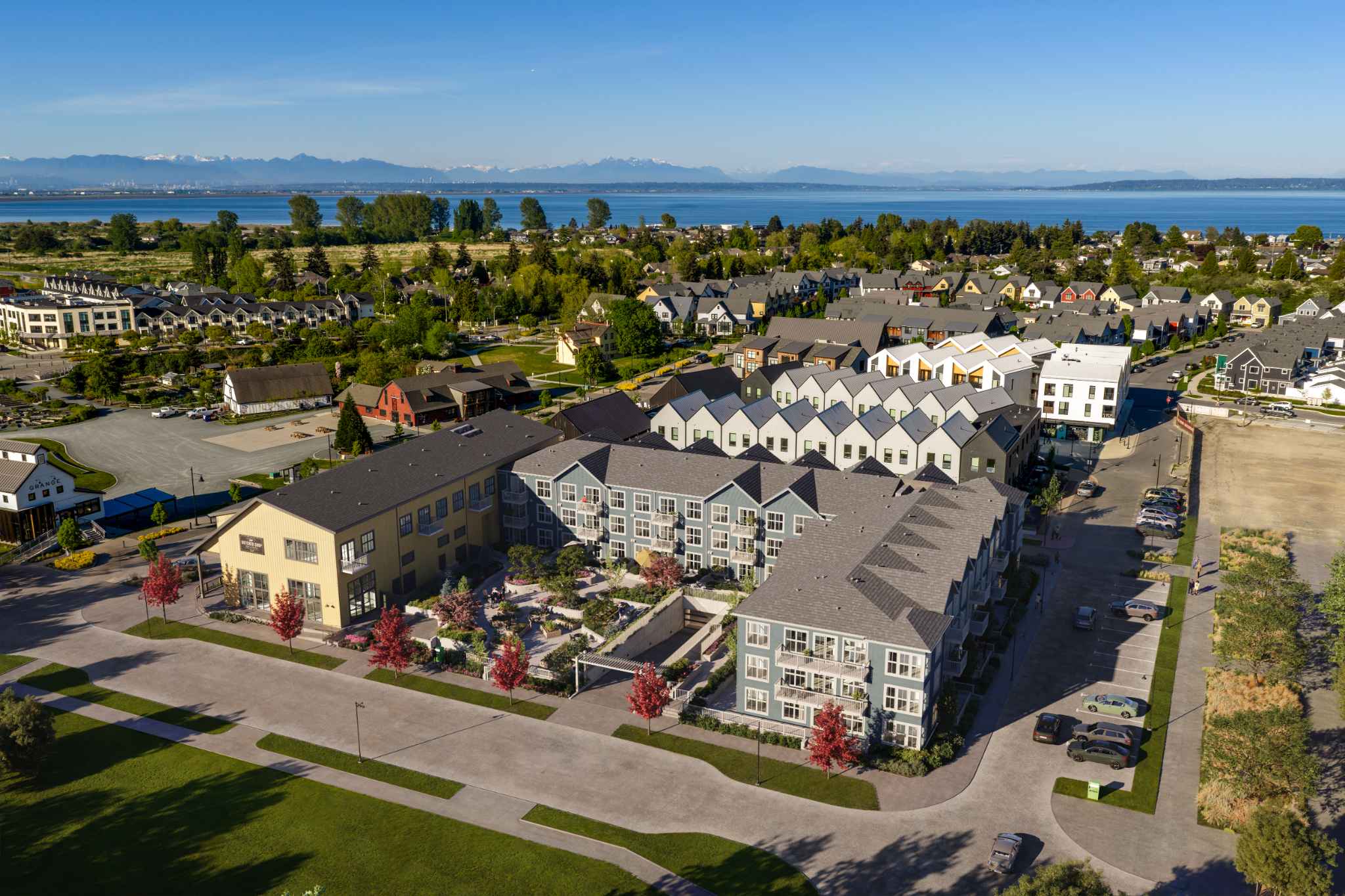 Market Commons – An elevated aerial view showcases a master-planned community of modern multi-family residences and gabled townhomes centered around an internal courtyard and parking area. In the background, green fields and residential neighborhoods stretch toward an expansive ocean bay and distant mountains under a clear blue sky.