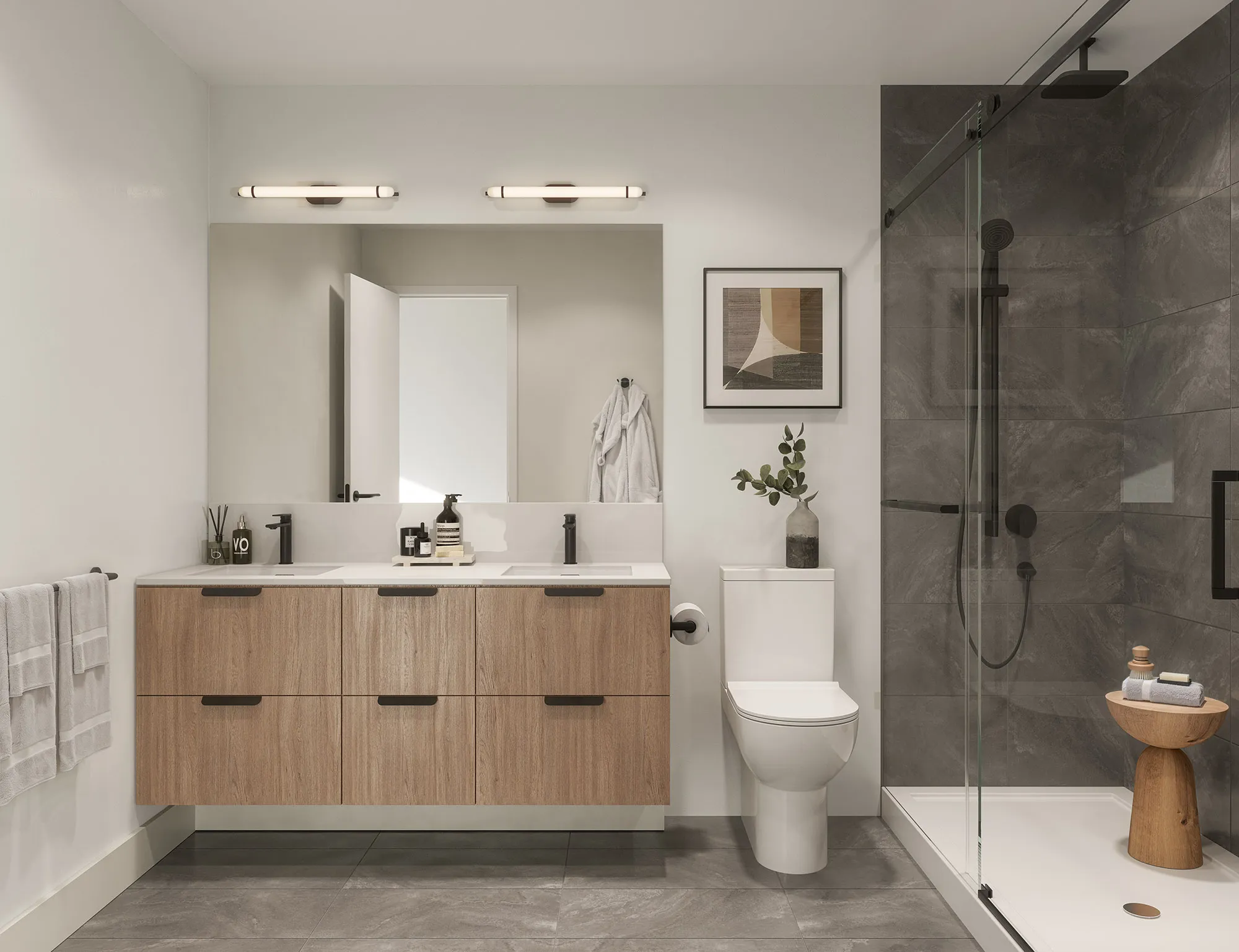 Level – A modern, bright bathroom featuring a floating light-wood double vanity with matte black fixtures, a white toilet, and a glass-enclosed walk-in shower tiled in dark grey stone. The space is illuminated by sleek wall sconces above a large frameless mirror and finished with large-format grey floor tiles.