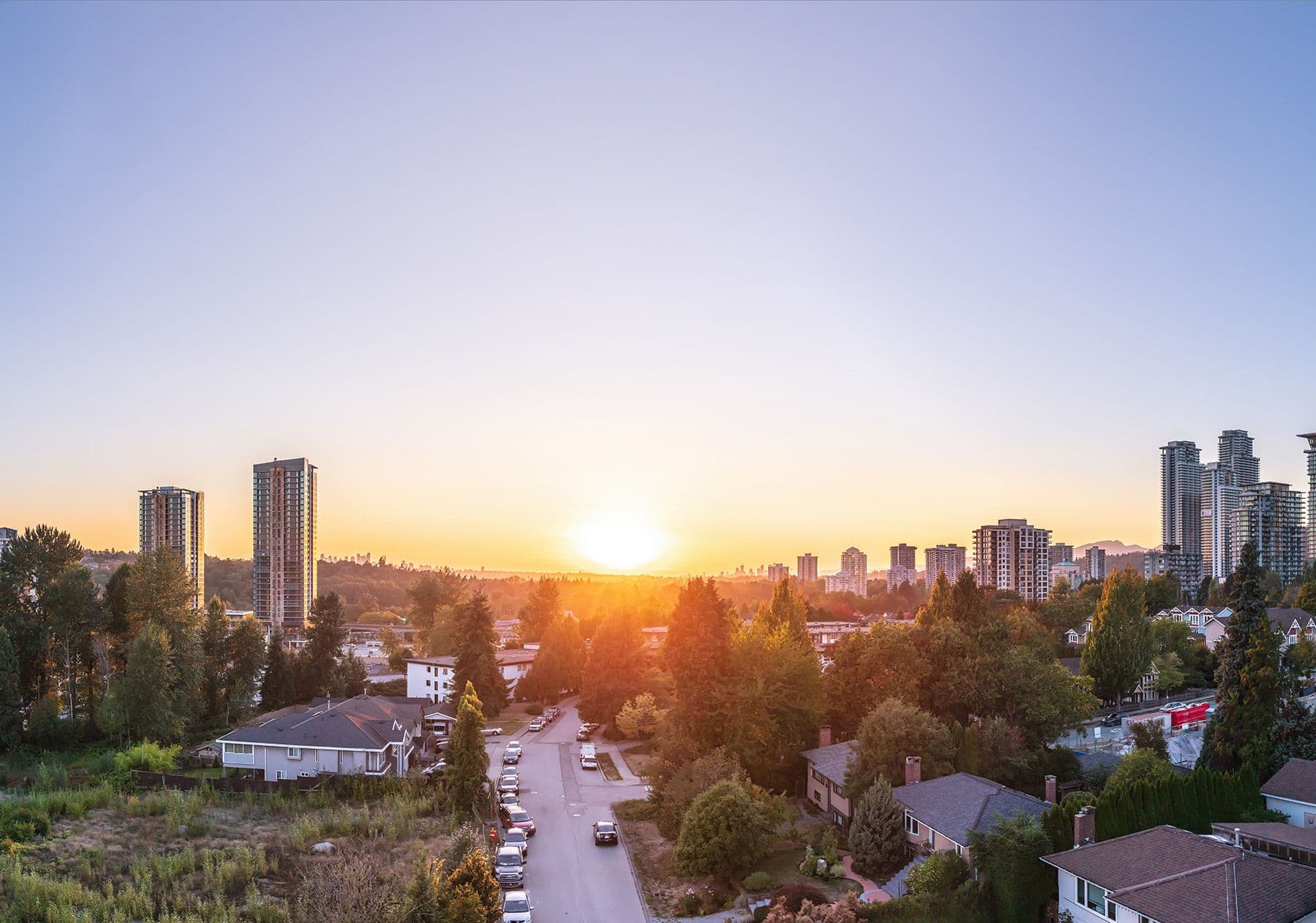 Level – A wide elevated view overlooks a quiet suburban street nestled among lush green trees and low-rise houses, flanked by modern residential high-rise towers. A bright sun sets directly on the distant horizon, casting a warm golden glow across a clear gradient sky.