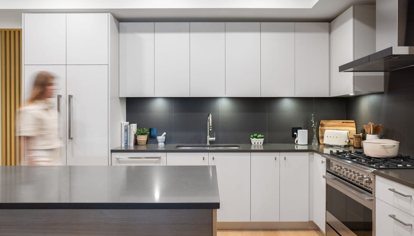 Bridge & Elliott – A modern, contemporary kitchen featuring clean white cabinetry paired with dark charcoal countertops and a matching dark backsplash. A large central island stands in the foreground, and high-end stainless steel appliances including a gas range and range hood complete the sleek, brightly lit space.