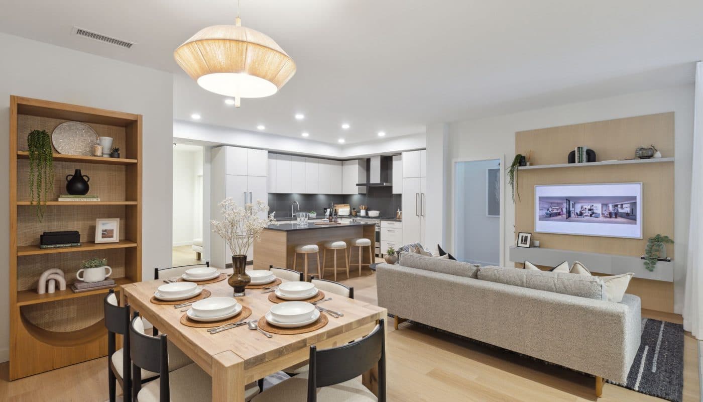 Bridge & Elliott – An open-concept living, dining, and kitchen area featuring light hardwood floors, clean white walls, and a warm wooden bookshelf. The space is furnished with a set wooden dining table, a grey sofa facing a wall-mounted TV on a light wood accent panel, and a modern kitchen with an island and stools in the background.