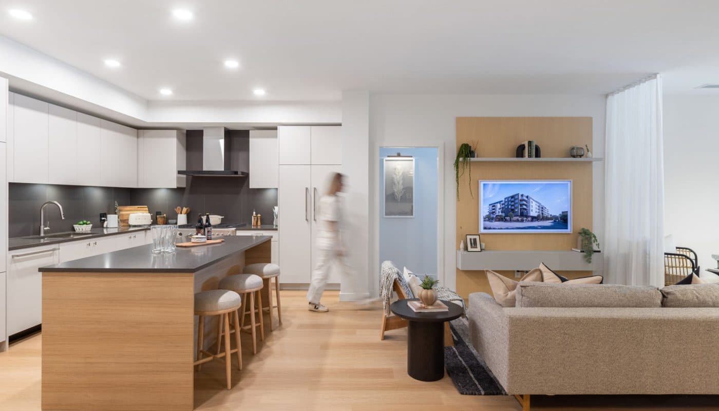 Bridge & Elliott – A bright, open-concept modern kitchen and living room featuring light wood flooring, a central island with barstools, and sleek white cabinetry with dark countertops. The living space includes a cozy neutral-toned sofa, a small coffee table, and a wall-mounted TV framed by a light wood accent panel.