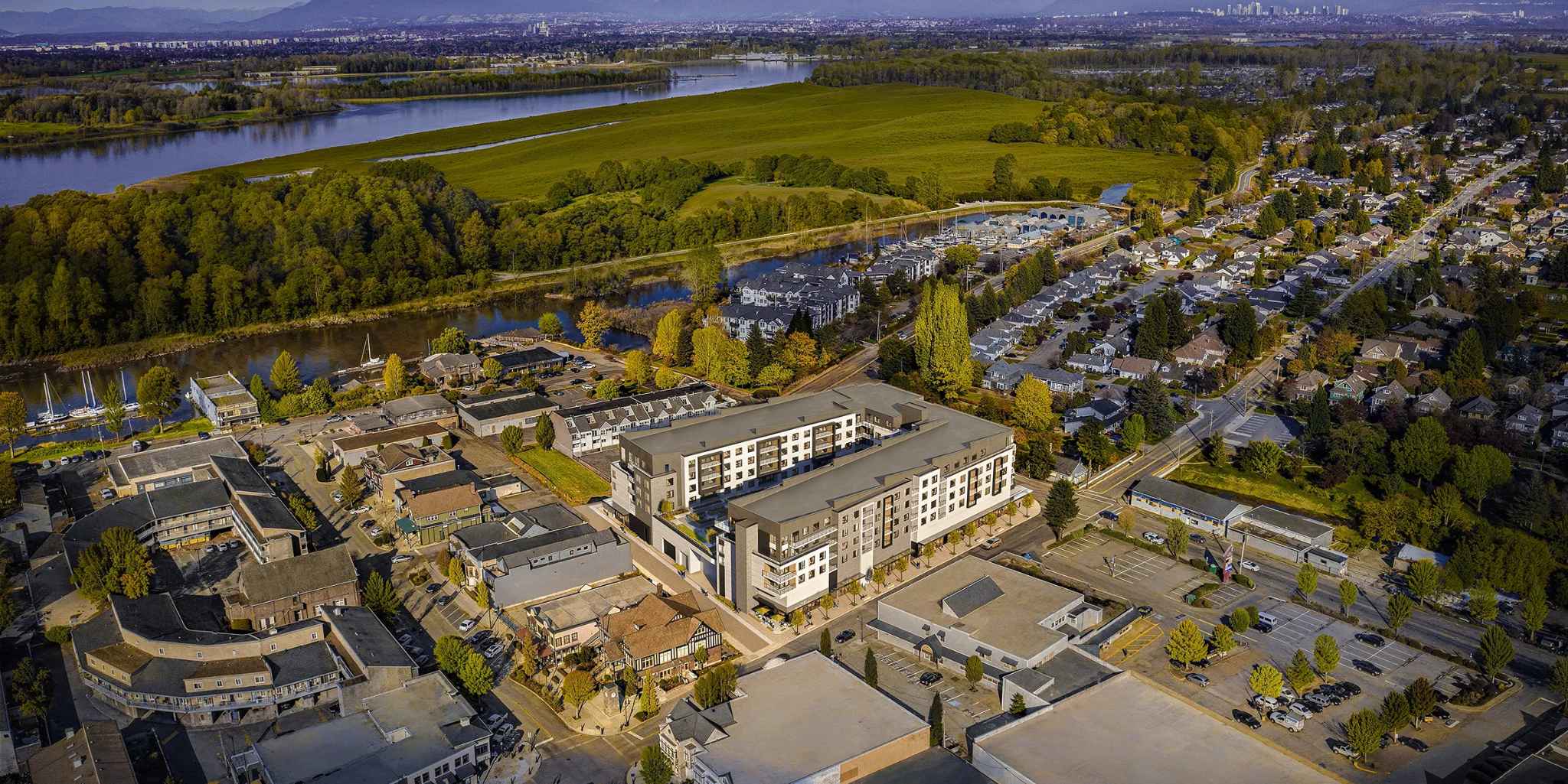 Bridge & Elliott – Aerial drone photo of a riverside town with a new mid-rise condo building highlighted among low-rise buildings, a marina, farmland, and river in the background.