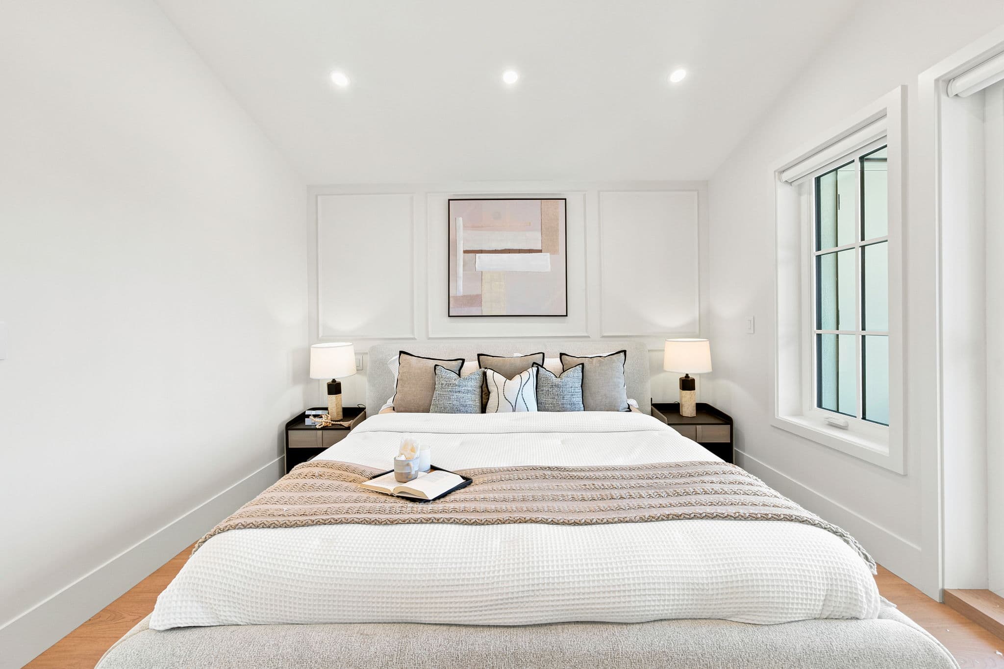 Carolin – A bright, symmetrical bedroom interior featuring a queen-sized bed dressed in white waffle-knit bedding, neutral accent pillows, and a textured throw with a reading tray. The feature wall showcases elegant picture frame molding, modern bedside tables with warm lamps, and a geometric abstract painting hung centered above the headboard.