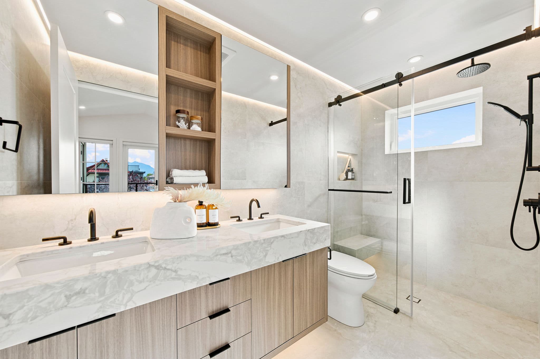 Carolin – A modern, bright ensuite bathroom featuring a floating light-wood double vanity with a thick marble countertop, matte black fixtures, and integrated under-cabinet lighting. Large-format beige tiles cover the walls and floor, extending into a glass-enclosed walk-in shower with a matte black rain showerhead and a built-in bench.