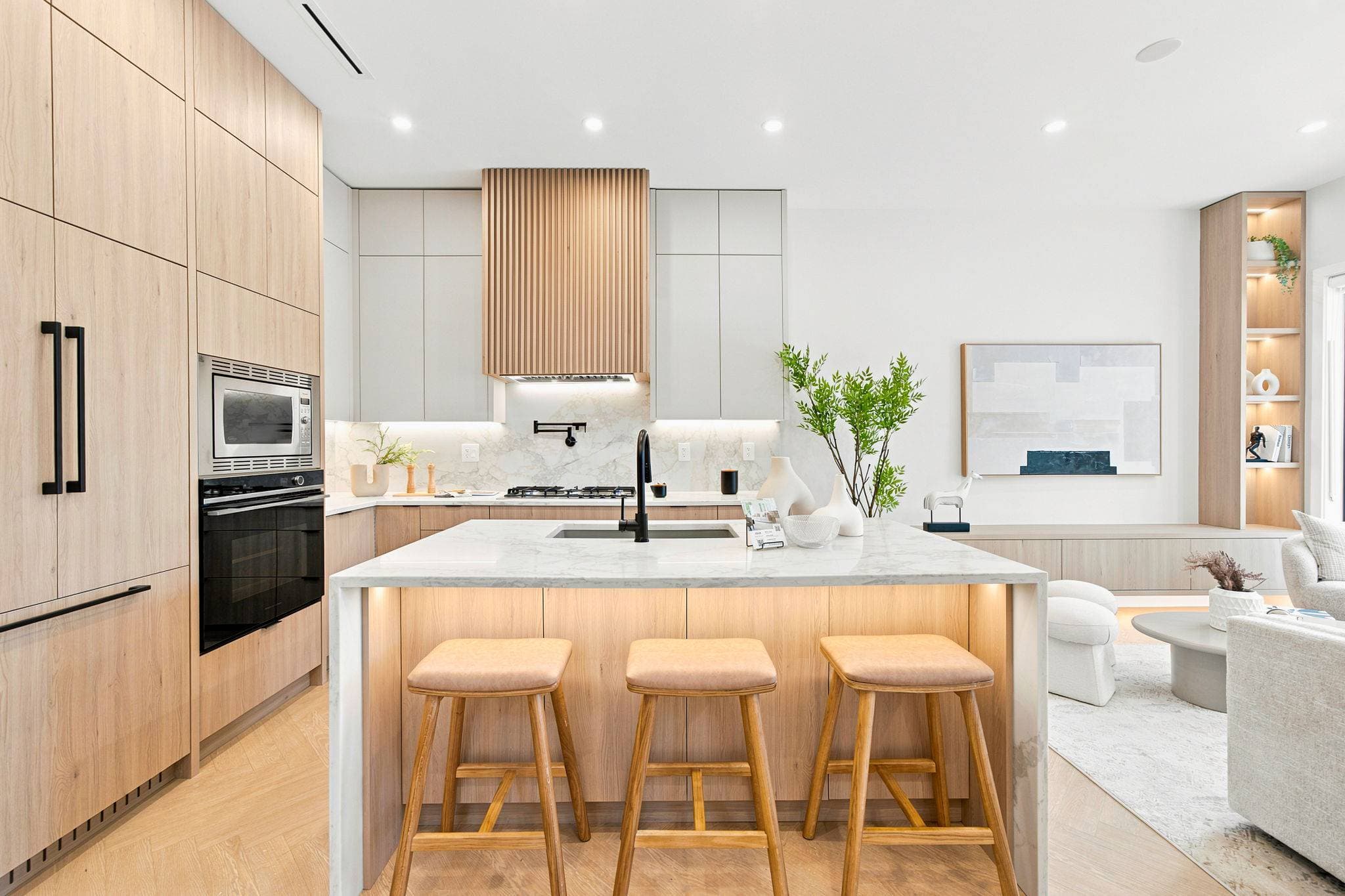 Carolin – A bright, modern kitchen featuring light wood cabinetry, integrated appliances, and a marble waterfall island with three barstools. Warm under-cabinet and under-counter lighting illuminates the space, which opens into an adjacent living area with soft neutral decor.