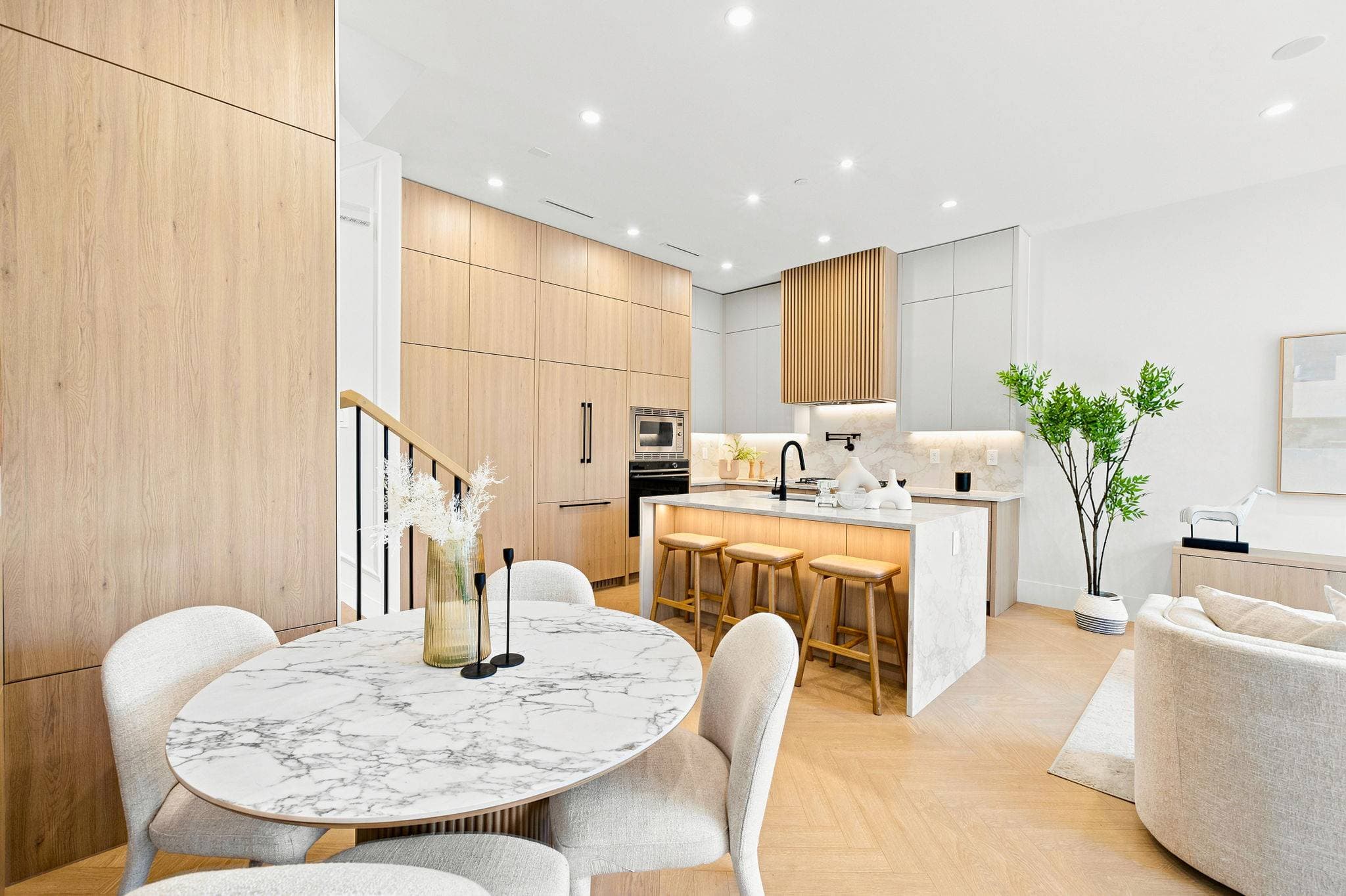 Carolin – A bright, open-concept kitchen and dining area featuring light natural wood cabinetry, a slatted range hood, and a marble waterfall island with barstools. In the foreground sits an oval marble dining table with upholstered chairs, set against light herringbone hardwood floors under warm recessed lighting.