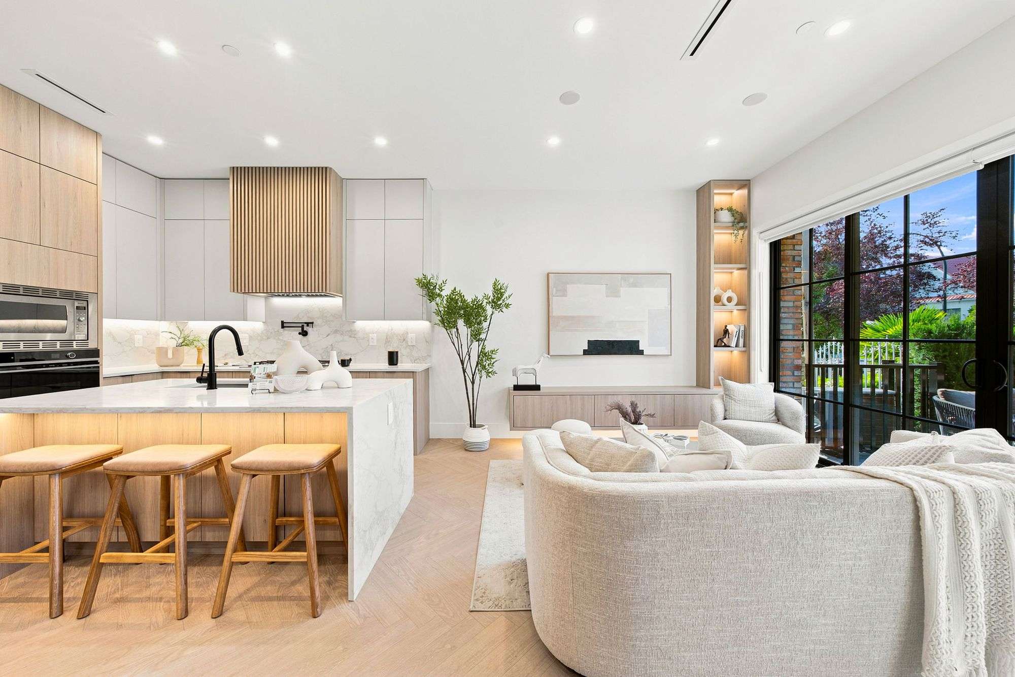 Carolin – A bright, open-concept living area and modern kitchen featuring light herringbone wood floors and neutral, contemporary furnishings. The kitchen showcases a marble waterfall island with wooden barstools, light wood and white cabinetry, and a slatted wood range hood, while large black-framed glass patio doors open to an outdoor space.