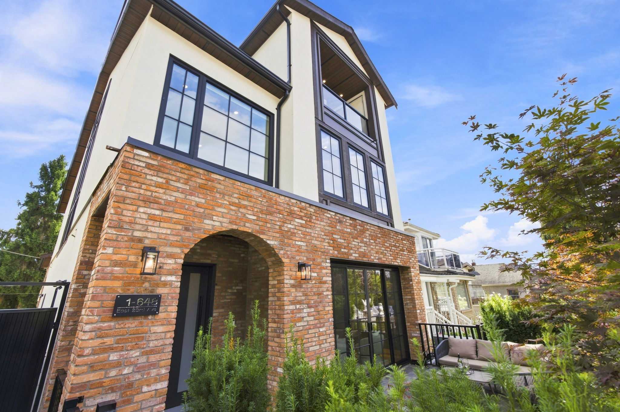 Carolin – A ground-level view of a contemporary multi-story home featuring a textured red brick facade on the lower level with an arched entryway and light stucco with dark-trimmed windows above. A small landscaped patio with outdoor seating borders the front entrance under a bright, sunny blue sky.