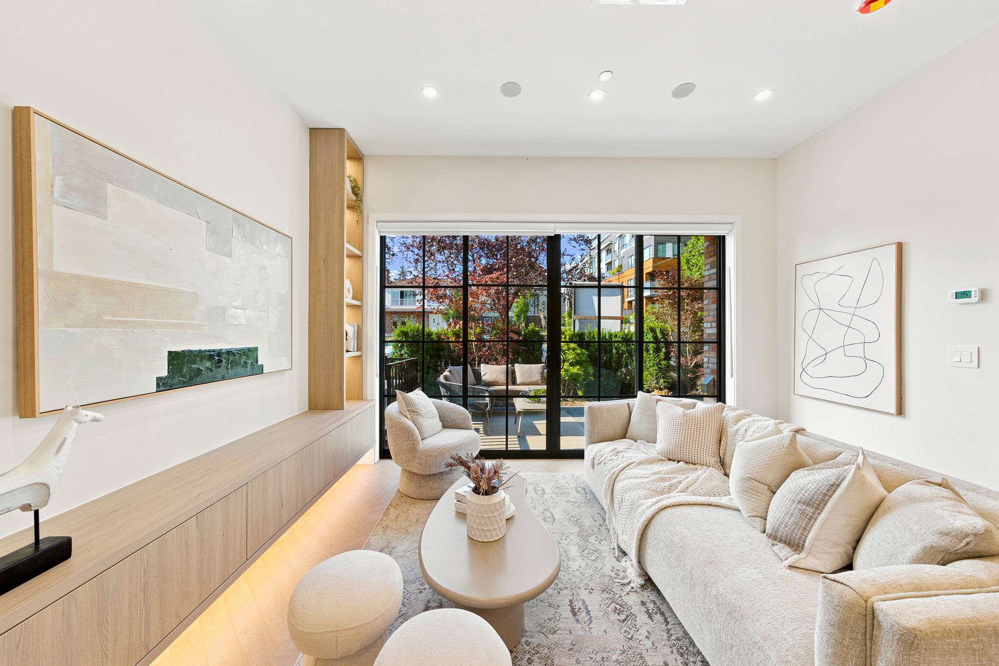 Carolin – A bright, contemporary living room features a plush cream sofa, curved armchair, and light wood built-in media console with soft under-cabinet lighting. Large black-framed French-style sliding glass doors open directly to a private outdoor patio framed by lush greenery.