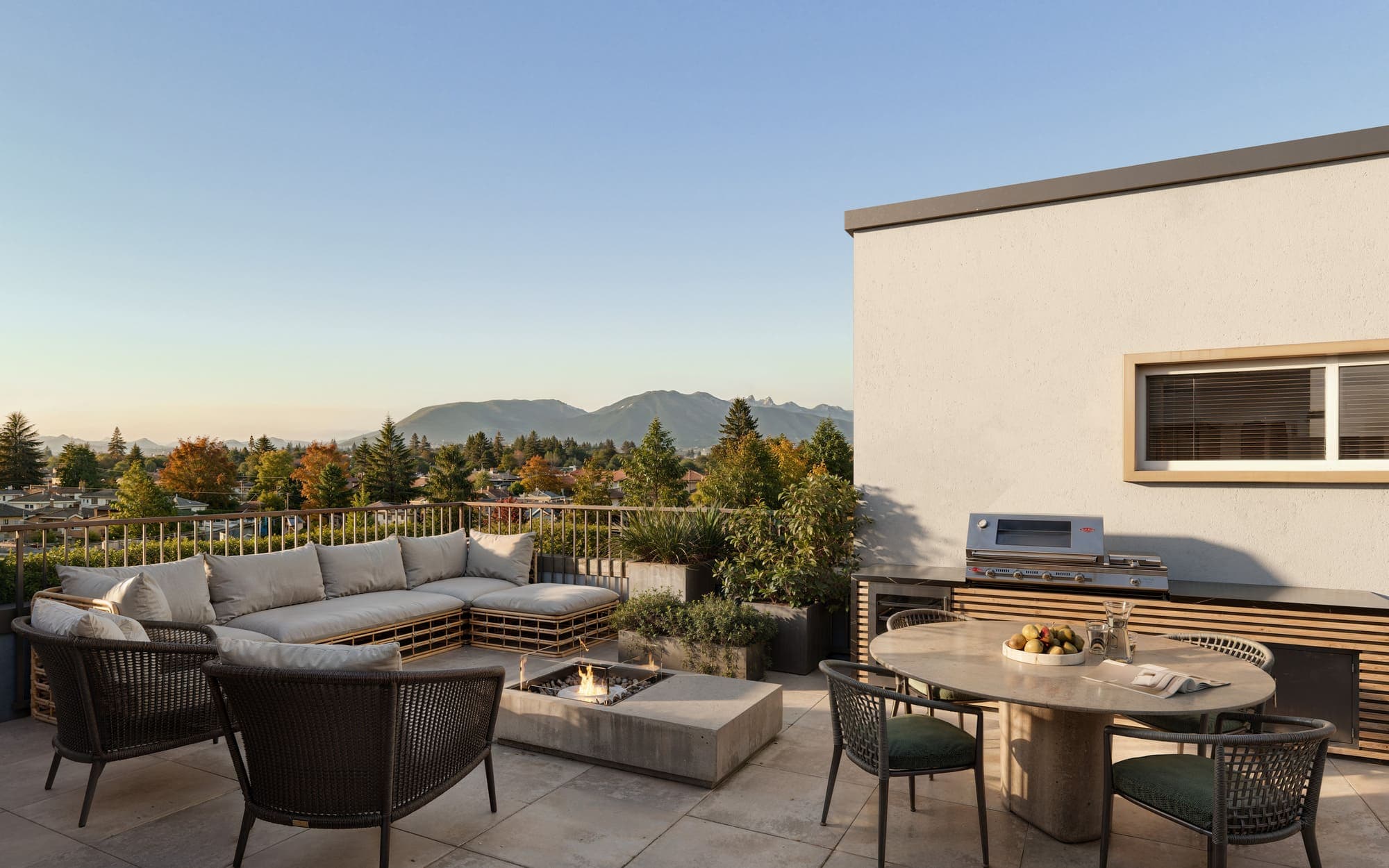 Oaklyn – A rooftop terrace with a sectional sofa, fire pit, dining table, and built-in BBQ station, overlooking the neighborhood and distant mountains.