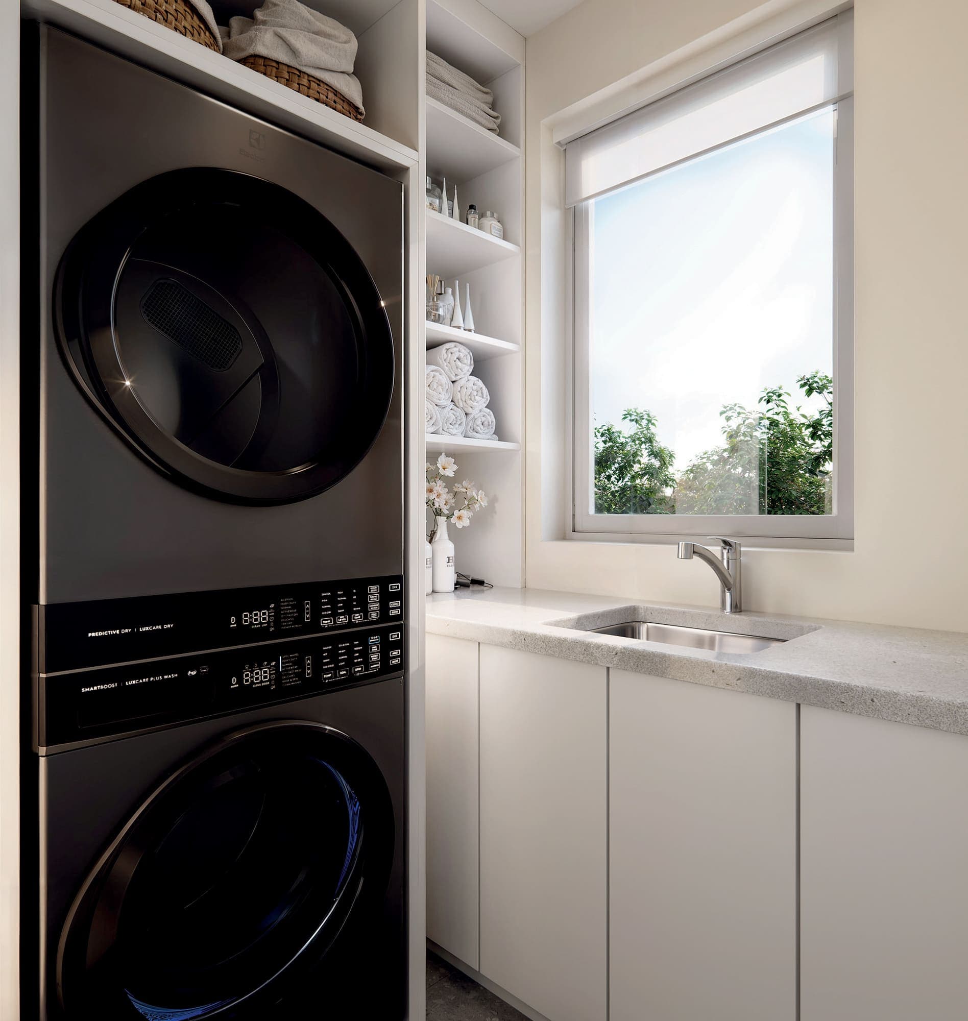 Oaklyn – A laundry nook with a stacked washer and dryer, open shelving with towels, and a small sink beneath a window looking out to greenery.