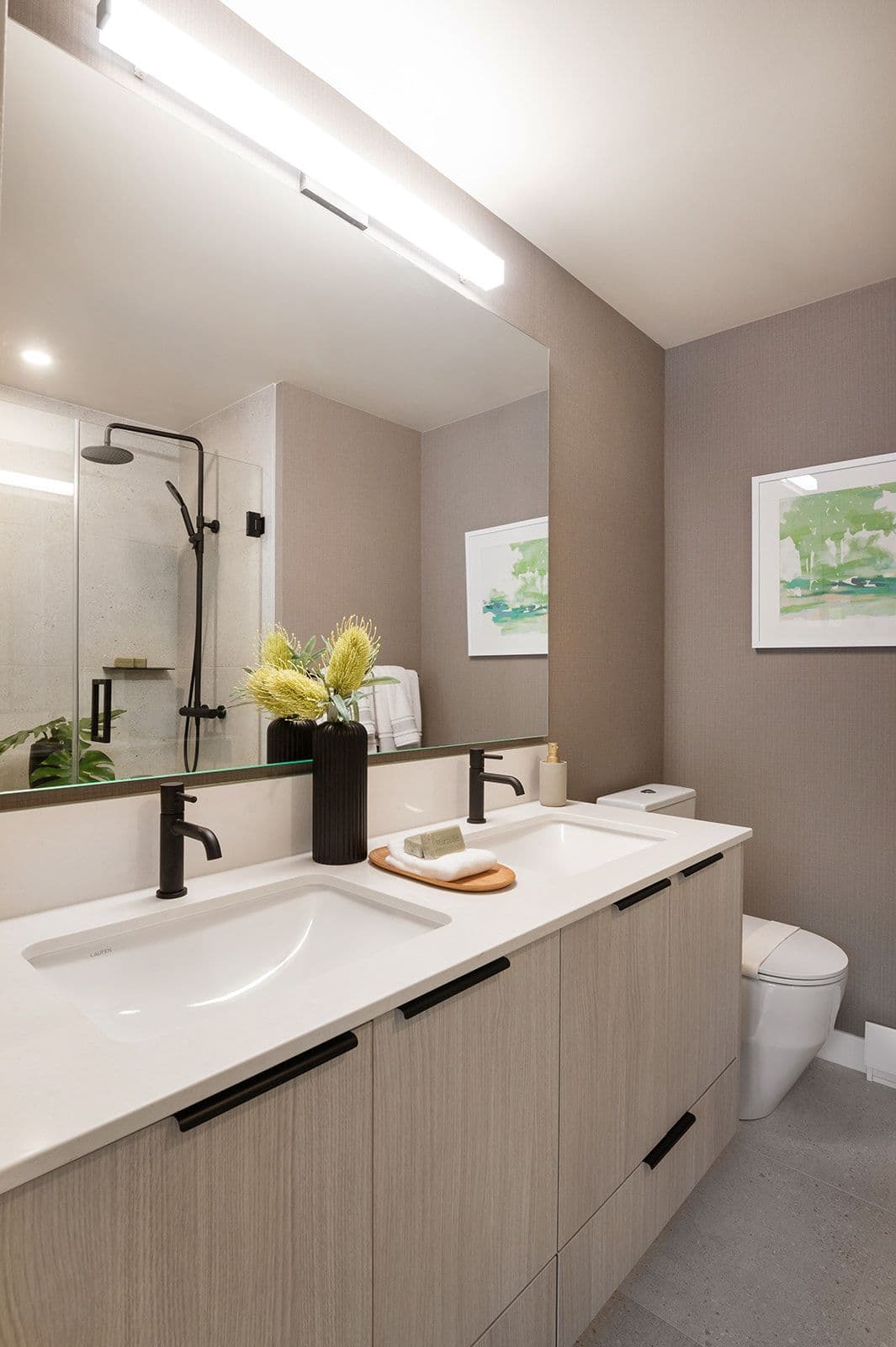 Sundays on Grant – A double-sink bathroom vanity with light wood cabinetry, matte black hardware, and a glass-enclosed shower in the reflection.