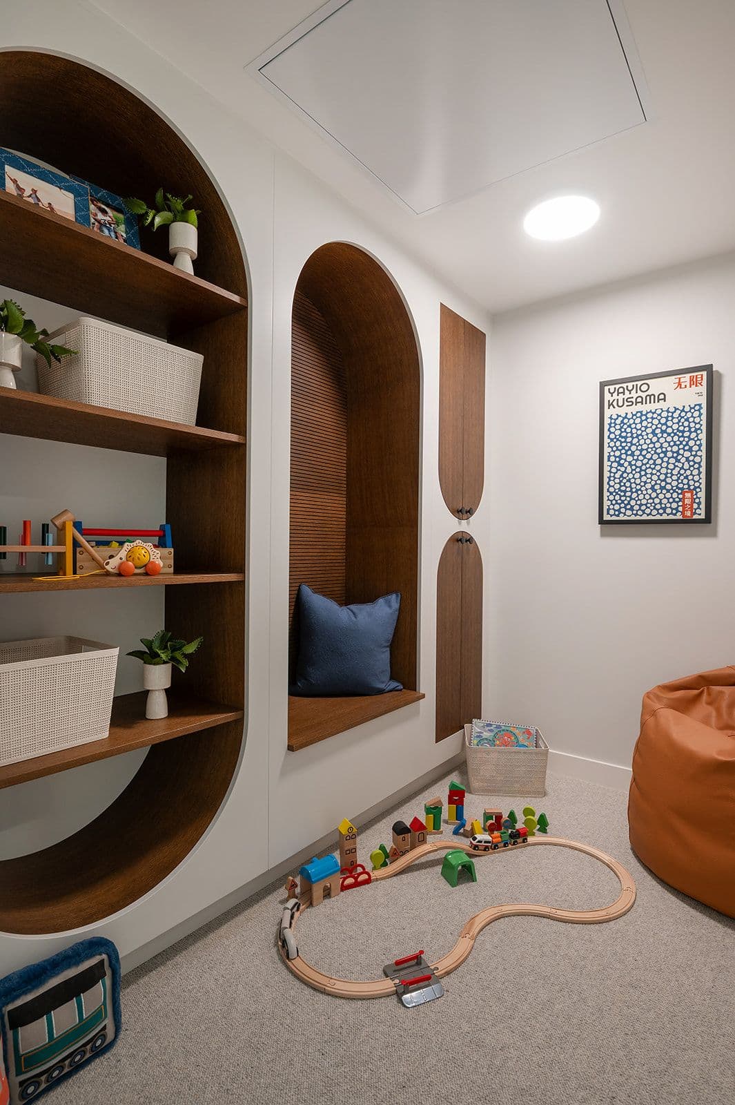 Sundays on Grant – A cozy children's playroom nook with arched built-in shelving, a seating alcove, and a wooden train set on carpeted flooring.