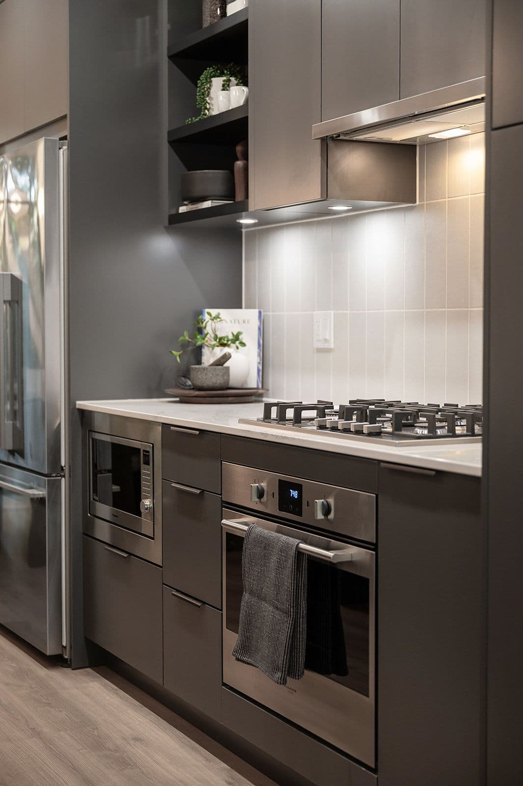 Sundays on Grant – Close-up of dark grey kitchen cabinets with built-in gas cooktop, oven, microwave, and under-cabinet tile lighting.