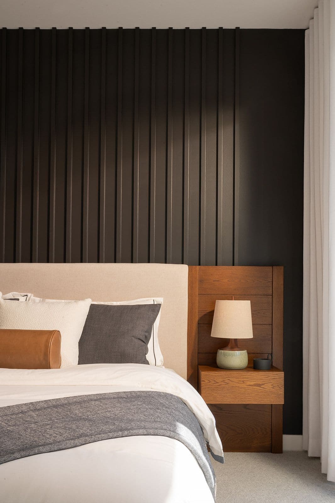 Sundays on Grant – Bedroom setup showing a plush upholstered headboard against a dark vertical slat wall and a wooden nightstand with lamp.