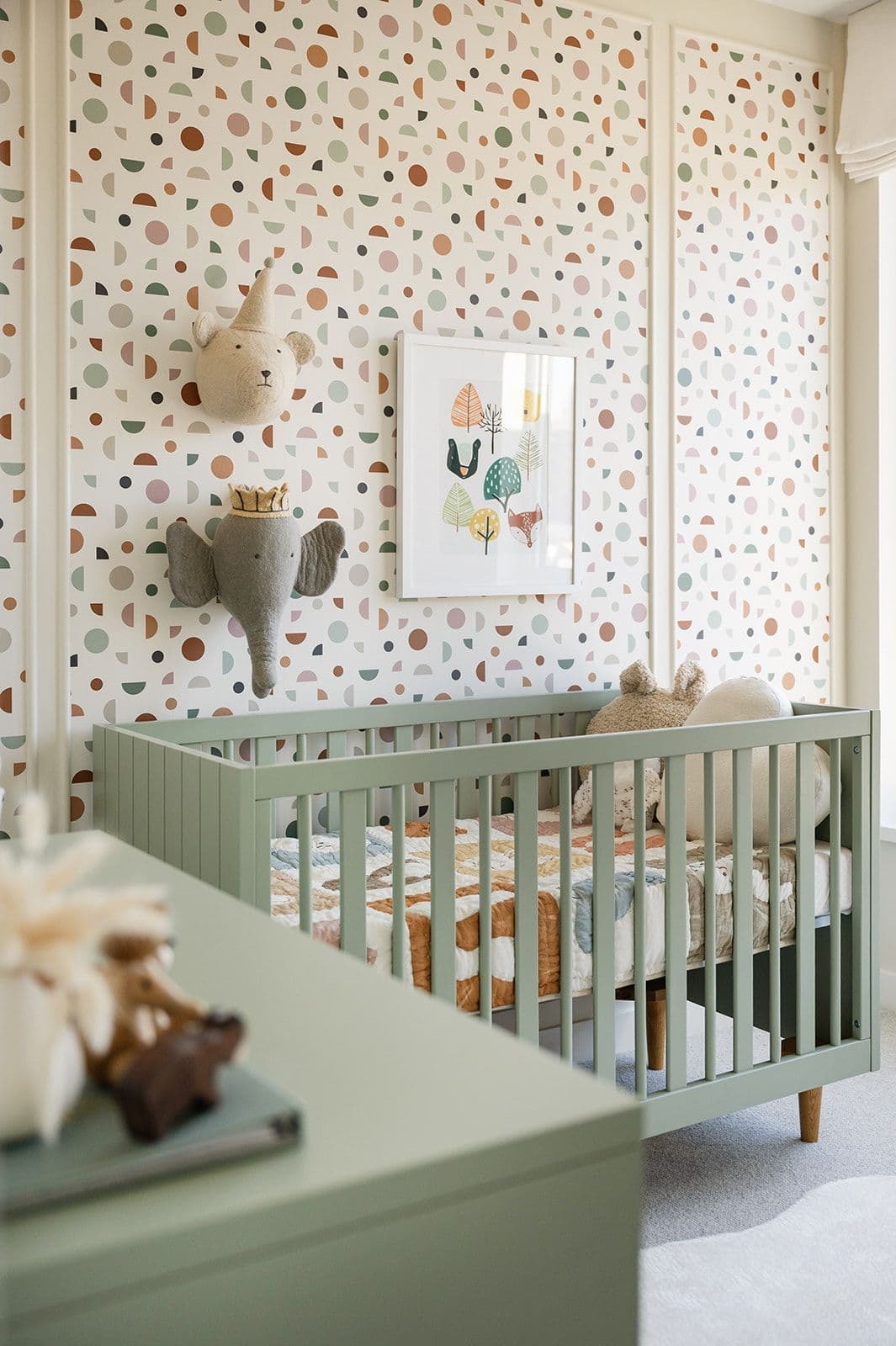 Sundays on Grant – A nursery bedroom with a sage green wooden crib, patterned geometric wallpaper, and plush animal wall decor.