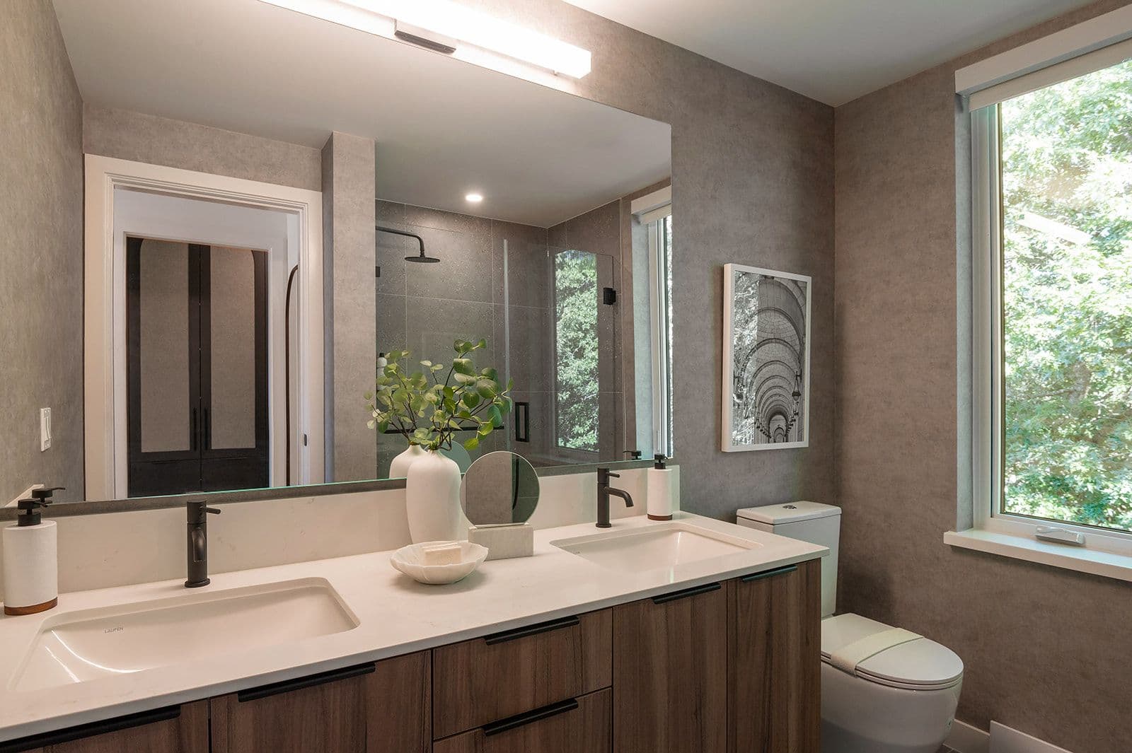 Sundays on Grant – Ensuite bathroom with dark wood double vanity, large frameless mirror, glass shower enclosure, and window looking out onto trees.