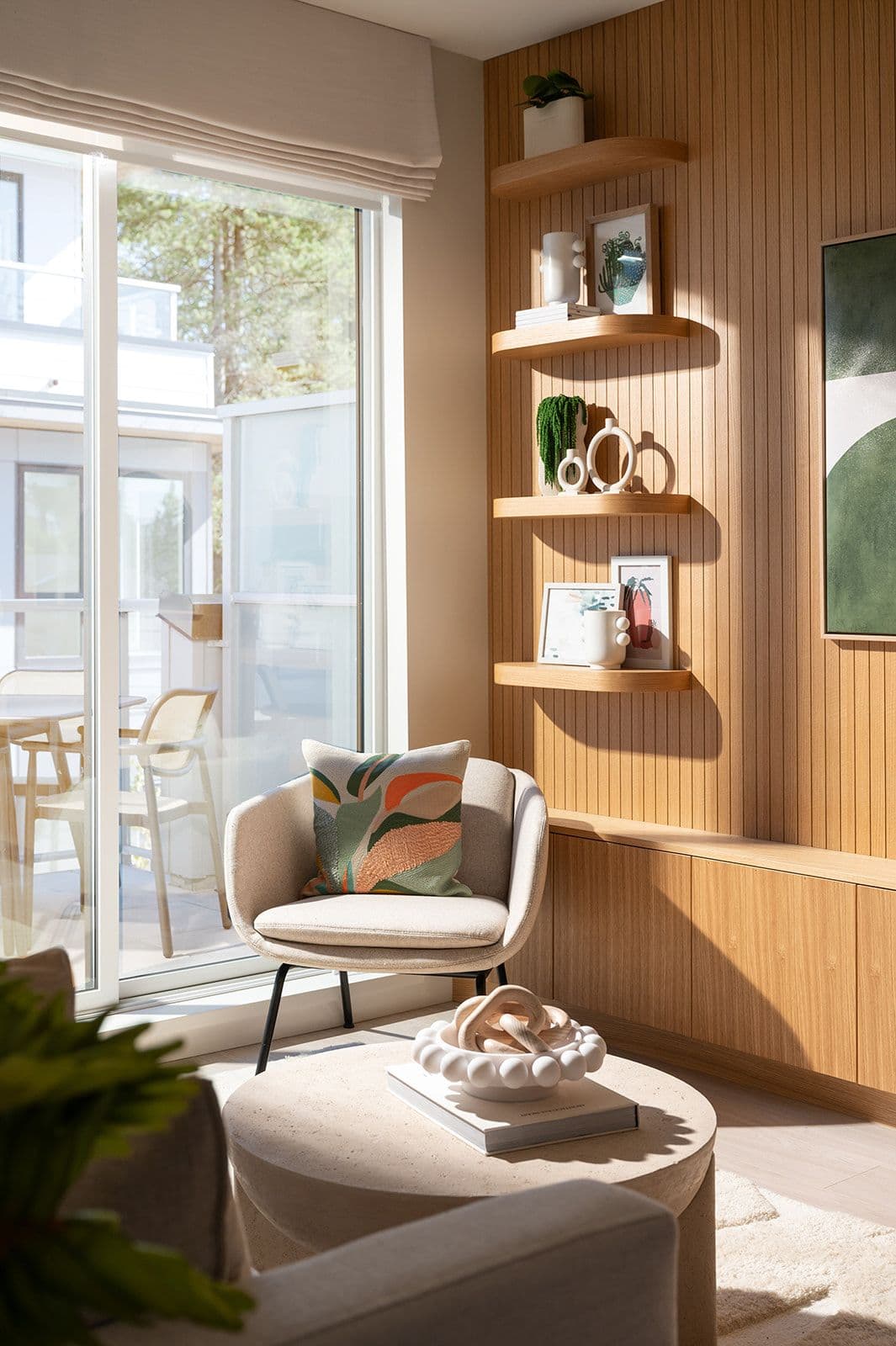 Sundays on Grant – Corner sitting area in a living room next to sliding glass balcony doors, featuring an armchair, floating wooden shelves, and slatted wall paneling.
