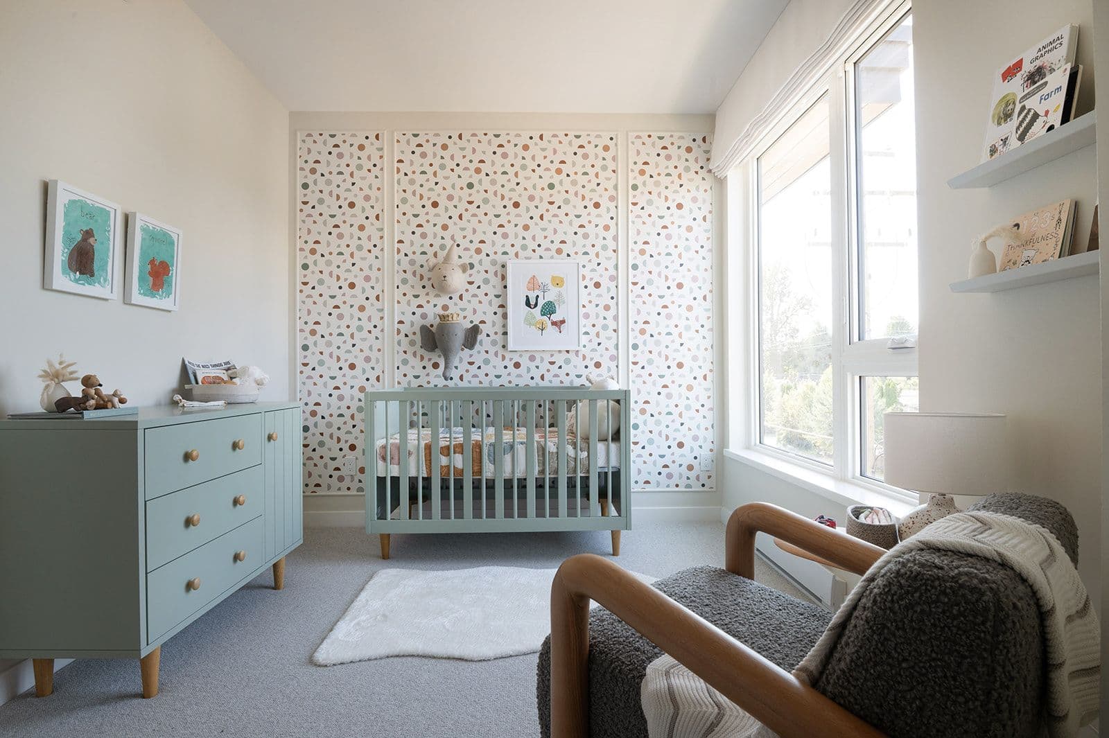 Sundays on Grant – Bright nursery room with a sage green crib, matching dresser, patterned accent wallpaper wall, and large window.