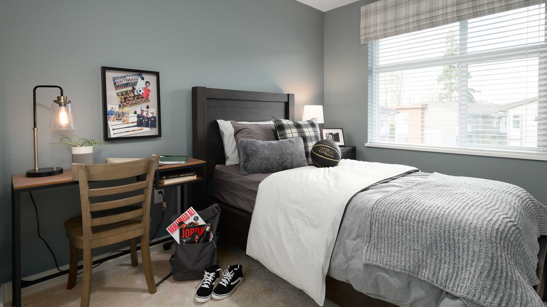 Aston – A boy's bedroom with a dark wood bed, sports-themed decor and photos, a desk with a lamp, and a large window.
