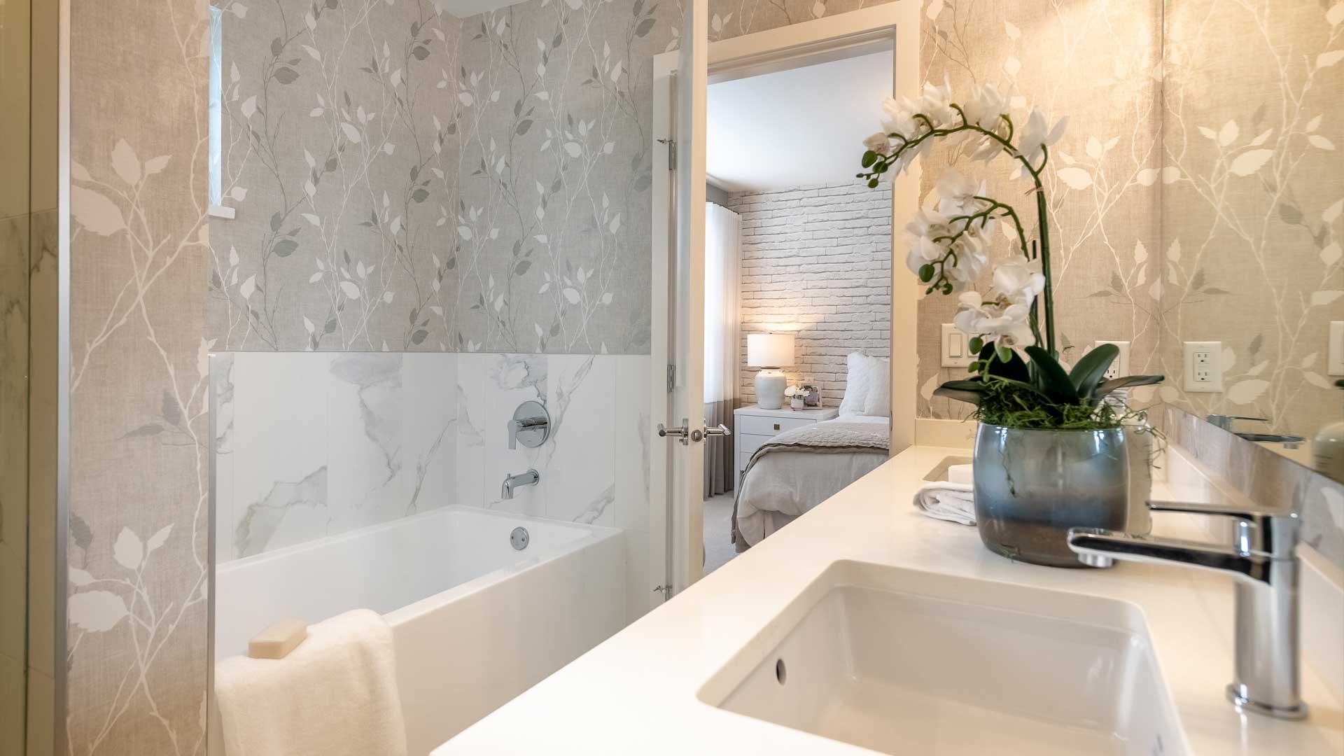 Aston – Bathroom with a soaking tub against floral wallpaper, a vanity with sink, and a doorway view into a bedroom with a brick accent wall.