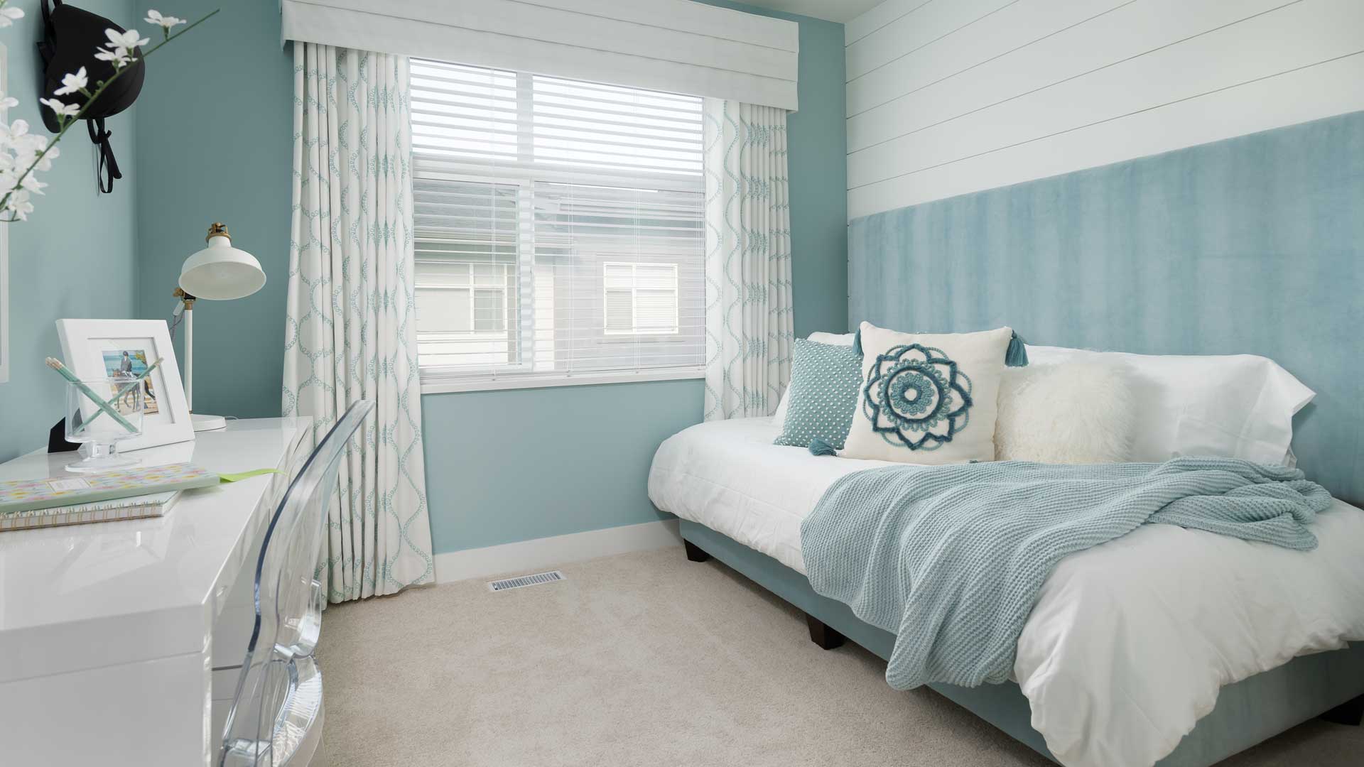 Aston – A girl's bedroom decorated in soft blue tones with a daybed, patterned pillows, curtains, and a white desk.