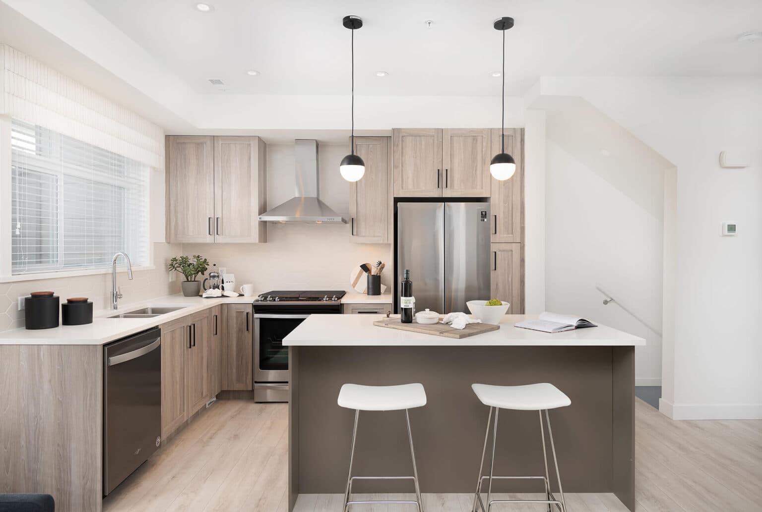 Aston – Kitchen with wood cabinetry, stainless steel appliances, a range hood, pendant lighting, and a white-topped island with bar stools.