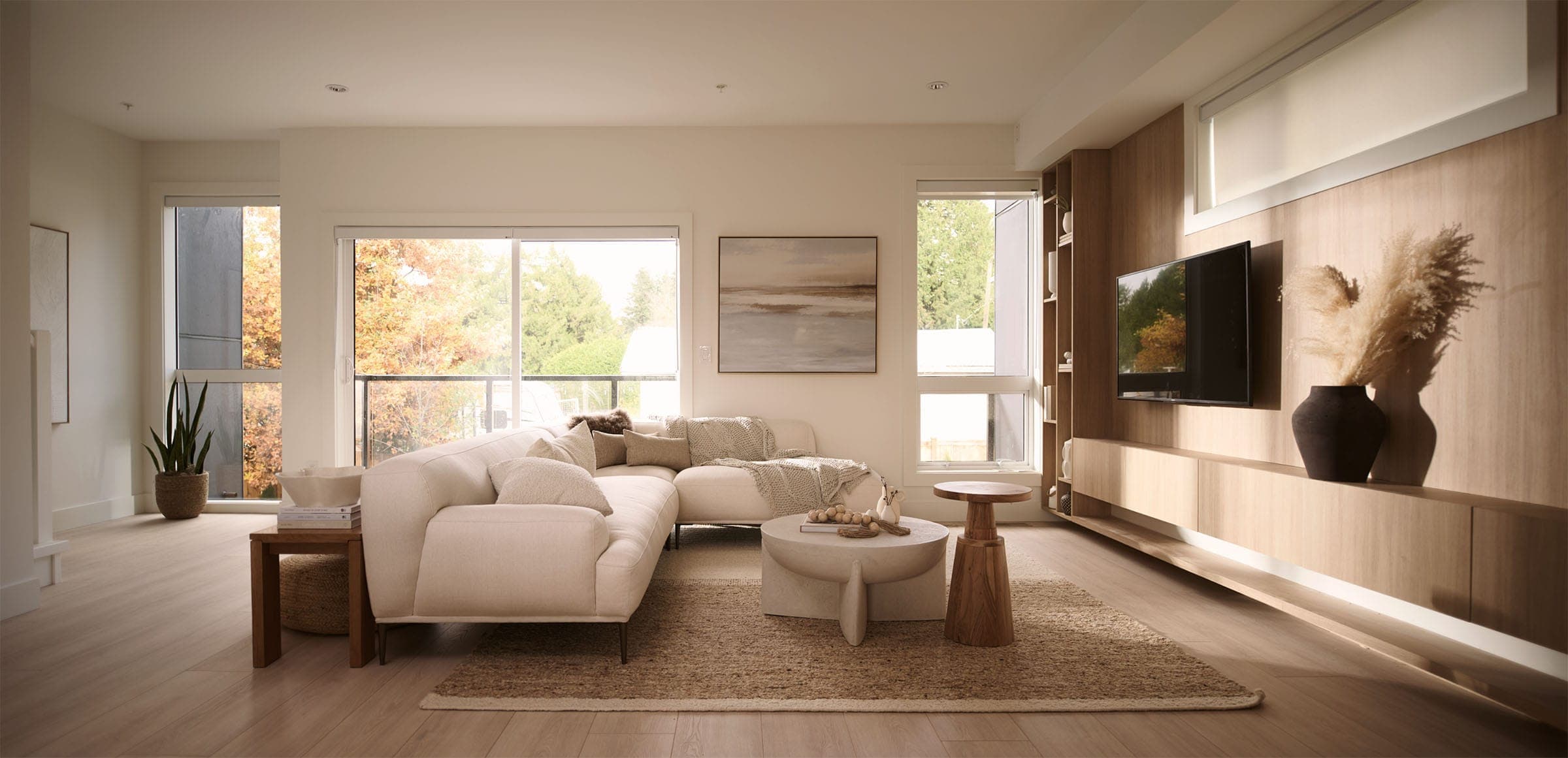 Addition Living – Living room with a large cream sectional sofa, a wood media wall with mounted TV, and floor-to-ceiling windows looking out to fall foliage.
