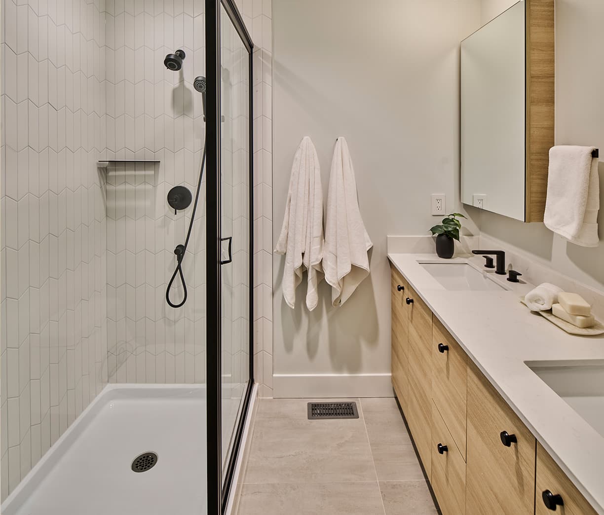 Addition Living – Bathroom close-up of a glass shower with vertical white tile and black fixtures, next to a light wood vanity with towels hanging on the wall.