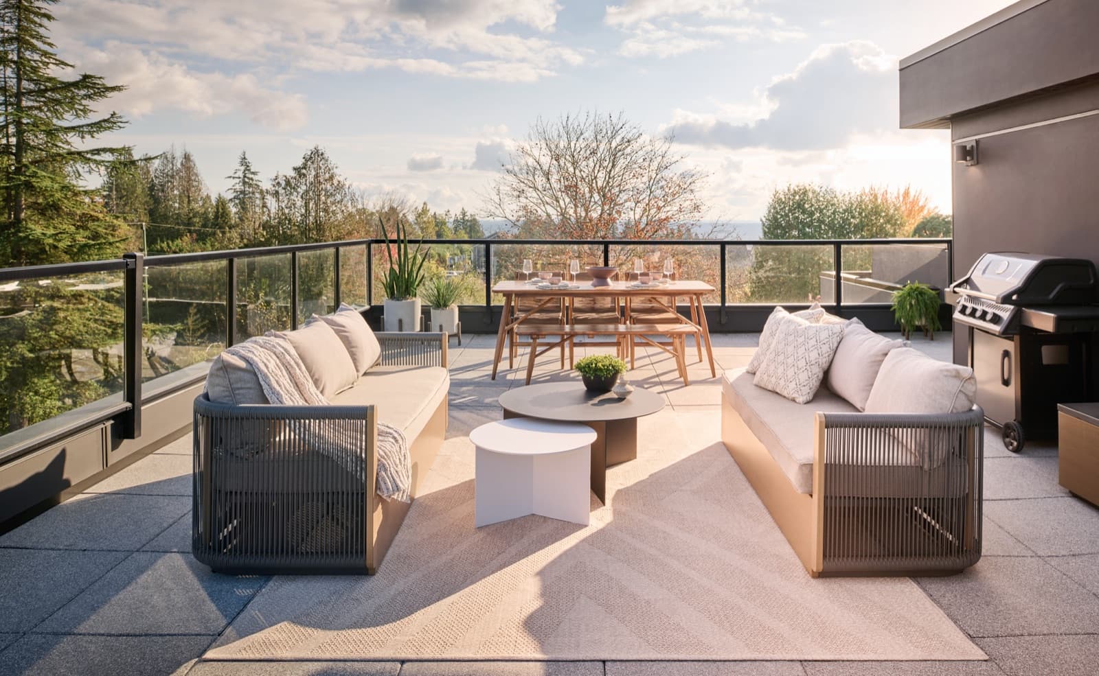 Addition Living – Rooftop deck with two outdoor sofas, a dining table set for six, and a built-in BBQ, overlooking trees and hills at sunset.