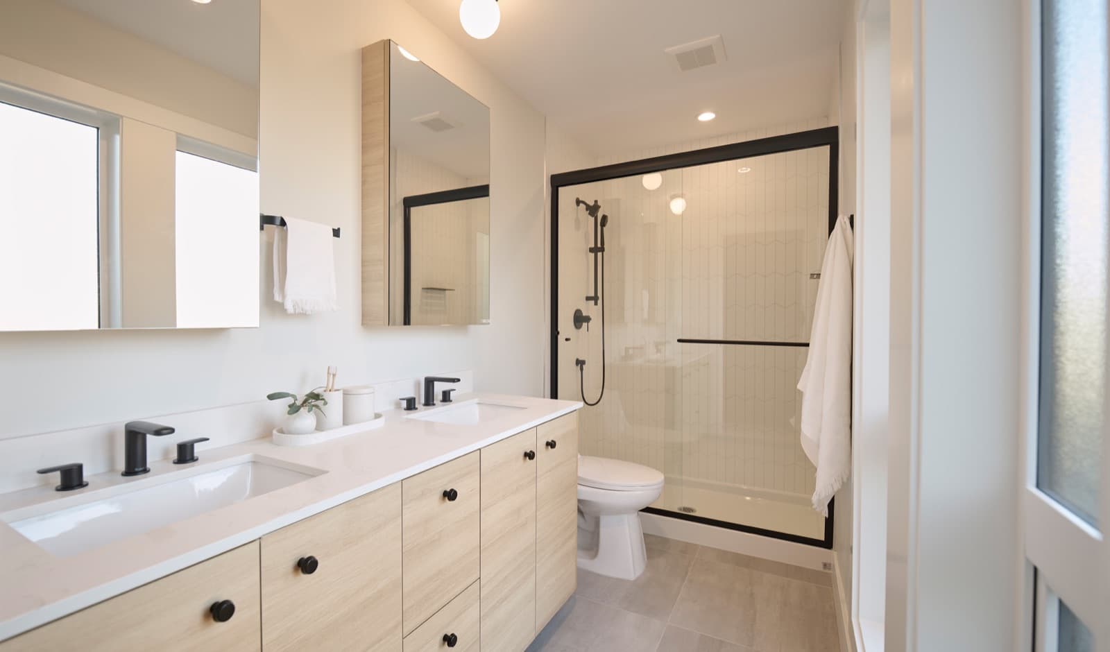 Addition Living – Bathroom with double sink vanity in light wood, black fixtures, and a glass-enclosed shower with white herringbone tile.