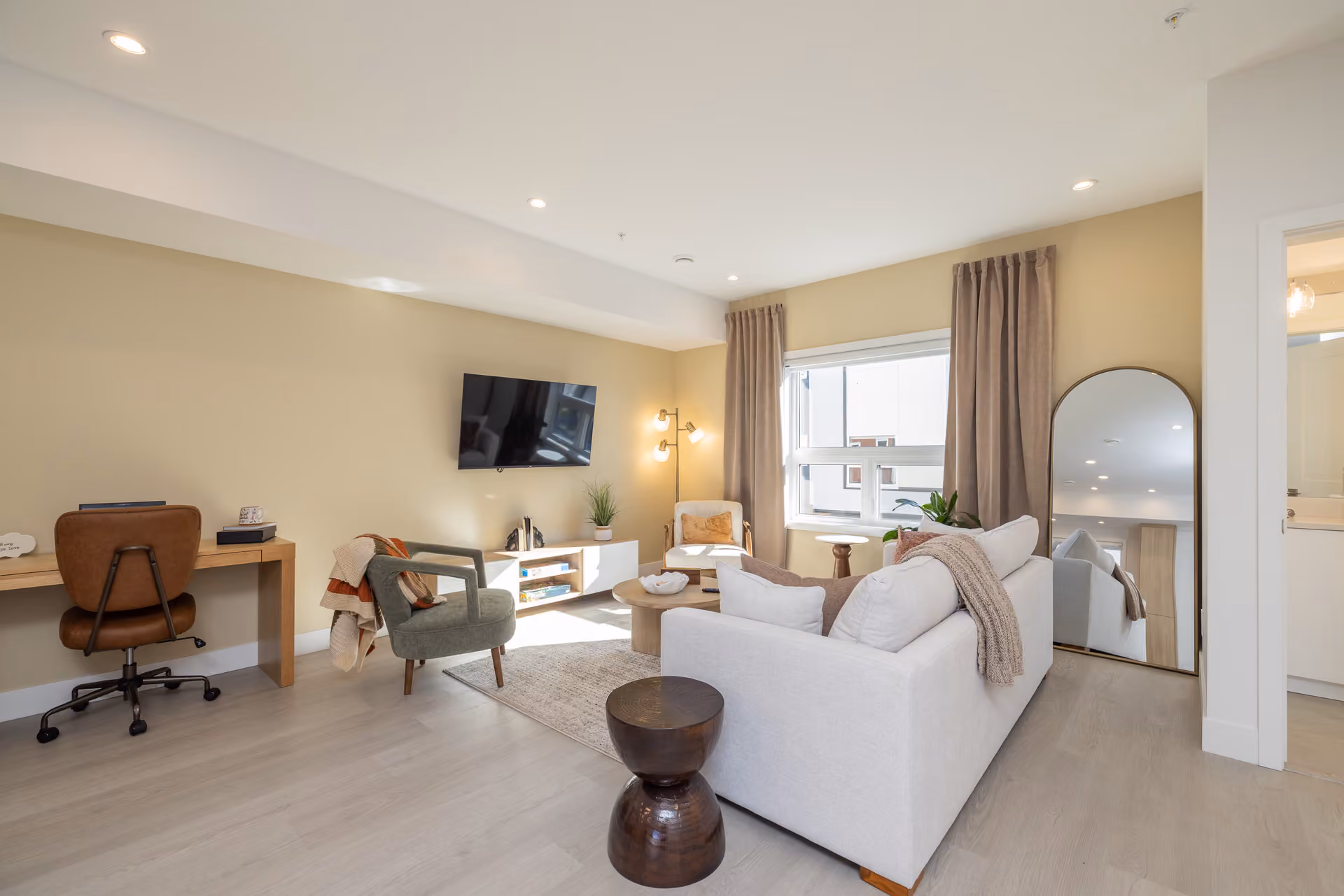 Skyview – A bright, open-concept living area featuring a white couch, a green armchair, and a dedicated desk space along light yellow walls. Warm daylight fills the room through a window framed by floor-length beige drapes, highlighting light wood flooring and modern accent furniture.