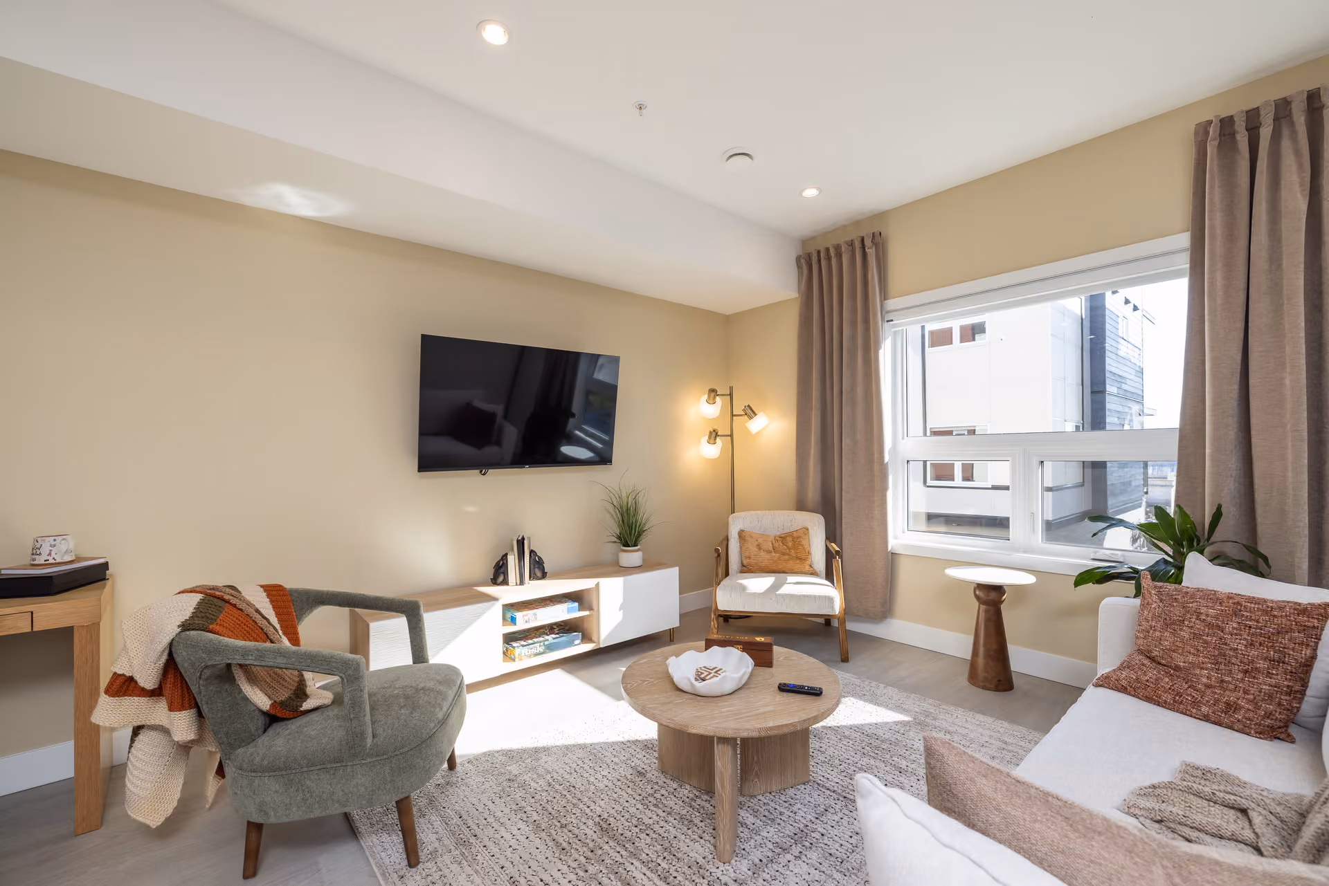 Skyview – A sunlit, cozy living room features a wall-mounted flat-screen television above a light wood console, flanked by an olive green armchair and a beige accent chair. Large windows draped with taupe curtains fill the space with natural light, highlighting warm beige walls, neutral area rugs, and a round wooden coffee table.
