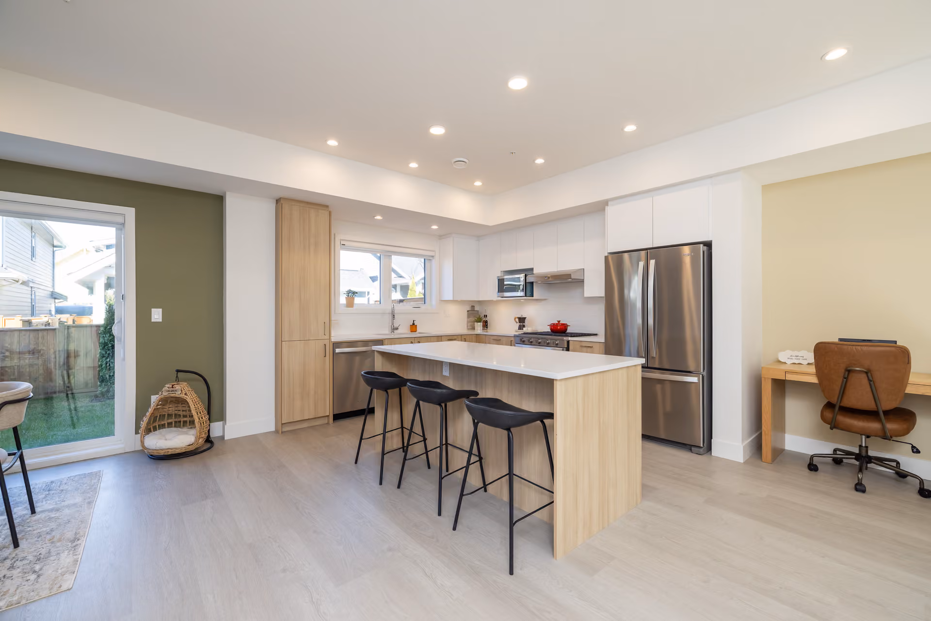 Skyview – A modern open-concept kitchen features light wood lower cabinets, white upper cabinets, stainless steel appliances, and a central island with three black barstools. Adjacent to the kitchen is a small home office nook with a brown leather chair, and sliding glass doors lead out to a fenced backyard.