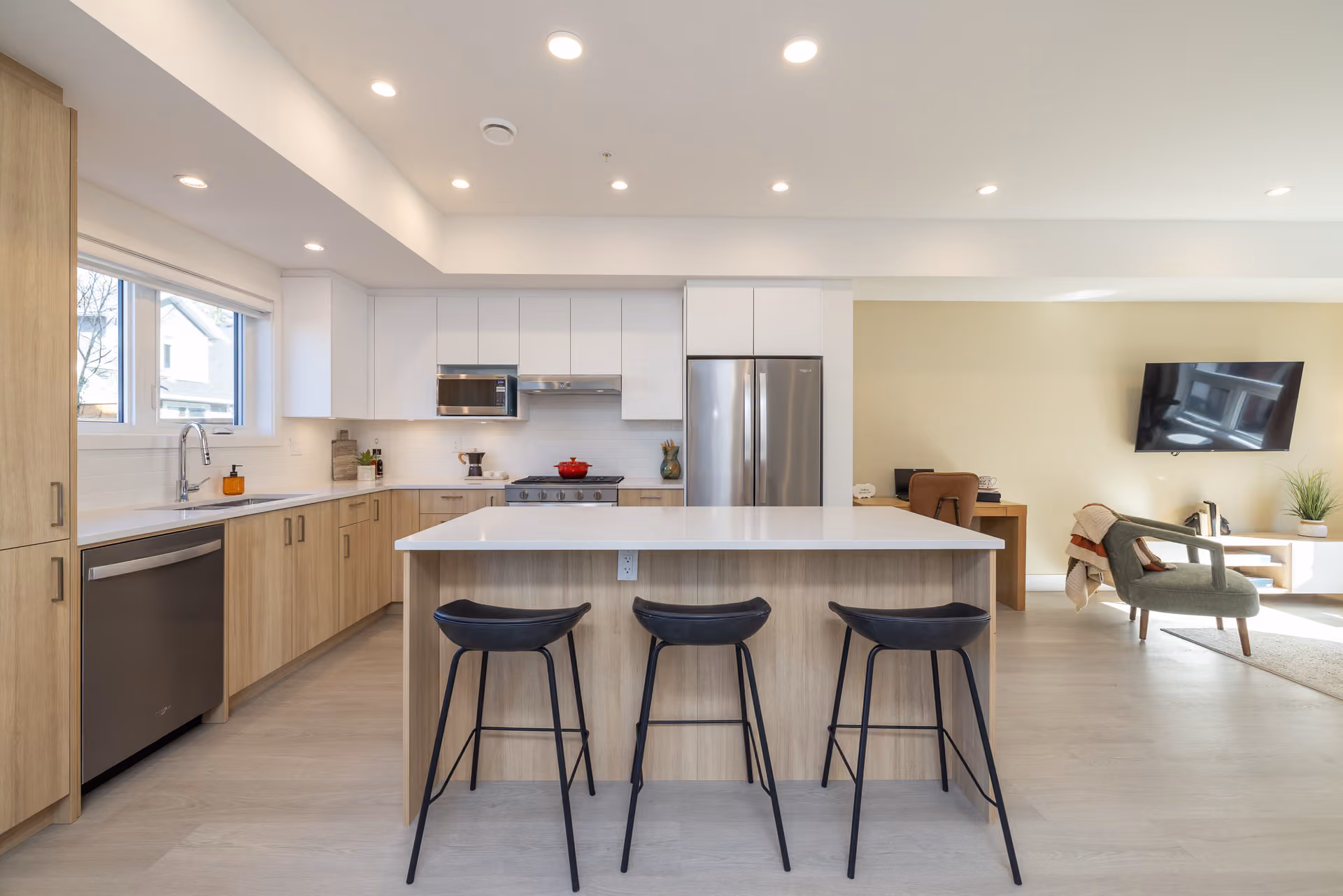 Skyview – A bright, open-concept modern kitchen features light wood lower cabinets, white upper cabinets, stainless steel appliances, and a central kitchen island with three black barstools. Light-colored hardwood flooring spans into the adjoining living area, which includes a wall-mounted television and a cozy armchair, all illuminated by recessed ceiling lights.