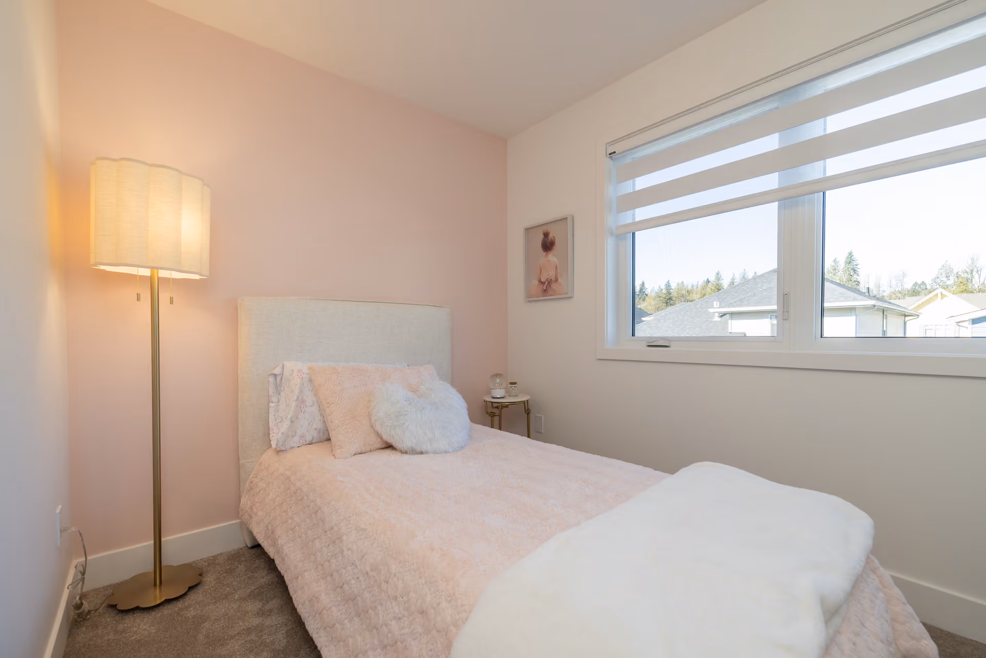 Skyview – A softly lit bedroom featuring a single bed with plush pink bedding, a heart-shaped pillow, and a pale upholstered headboard set against a blush pink accent wall. A tall standing lamp with a pleated shade glows beside the bed, while natural light streams in through a large window with modern horizontal zebra blinds.