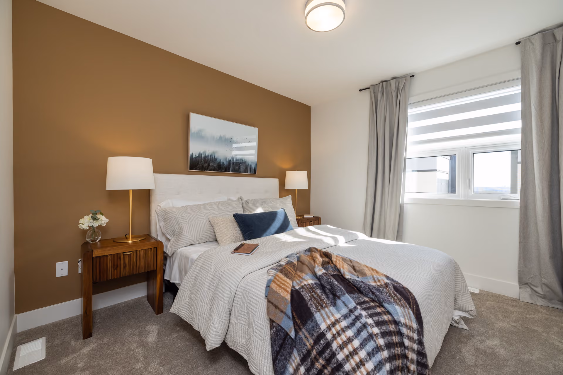 Skyview – A modern bedroom featuring a bed layered with grey patterned bedding, a plaid throw, and dark blue accent pillows against a warm ochre accent wall. Wooden nightstands with gold lamps flank the bed, while natural light streams in from a side window draped with full-length grey curtains.
