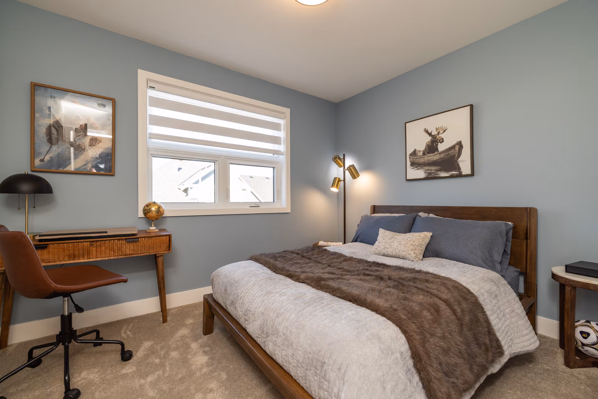 Skyview – A cozy bedroom with dusty blue walls featuring a wooden platform bed made with blue and grey bedding and a faux-fur throw. Next to the bed sits a mid-century wooden writing desk with a leather office chair, illuminated by light coming through a window with white zebra blinds.