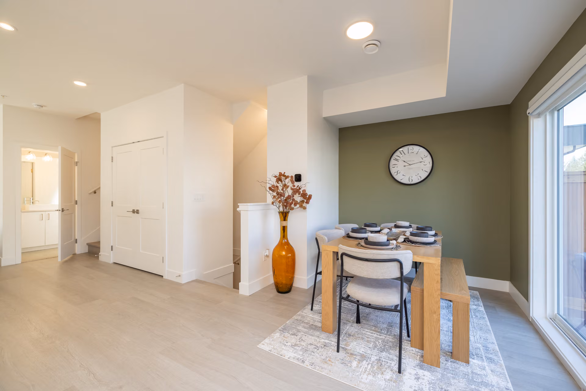 Skyview – A modern open-concept dining area featuring a wooden table set for a meal, a bench, and contemporary chairs against a dark olive-green accent wall adorned with a large round clock. Light wood floors extend throughout the space, which opens to a hallway with white double doors, a staircase, and a doorway leading into a brightly lit bathroom.