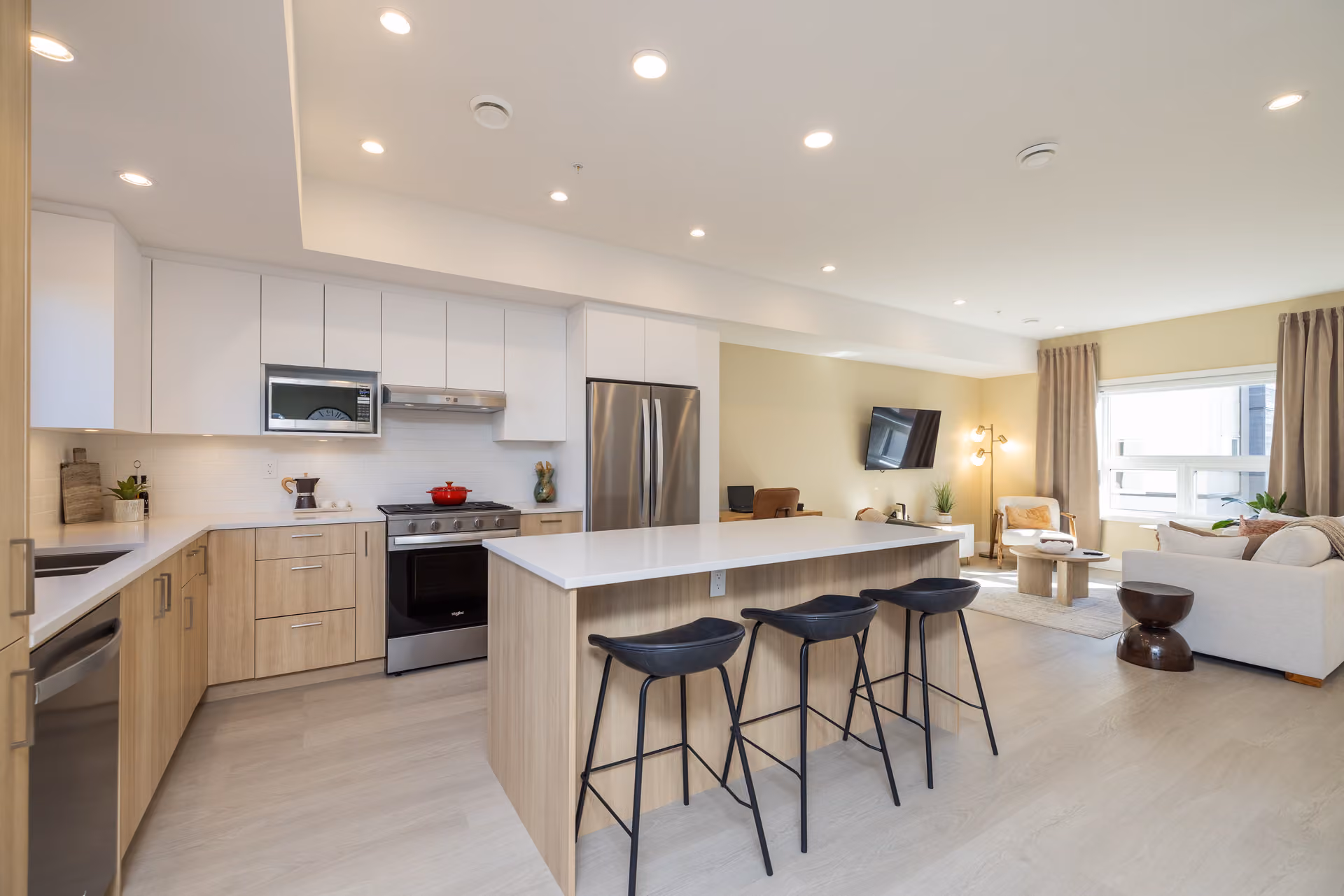 Skyview – An open-concept kitchen and living room featuring light wood low cabinets, flat white upper cabinets, stainless steel appliances, and a large central island with three black barstools. Natural light streams into the adjoining living area through a window at the back, illuminating soft neutral furnishings and light laminate flooring throughout.